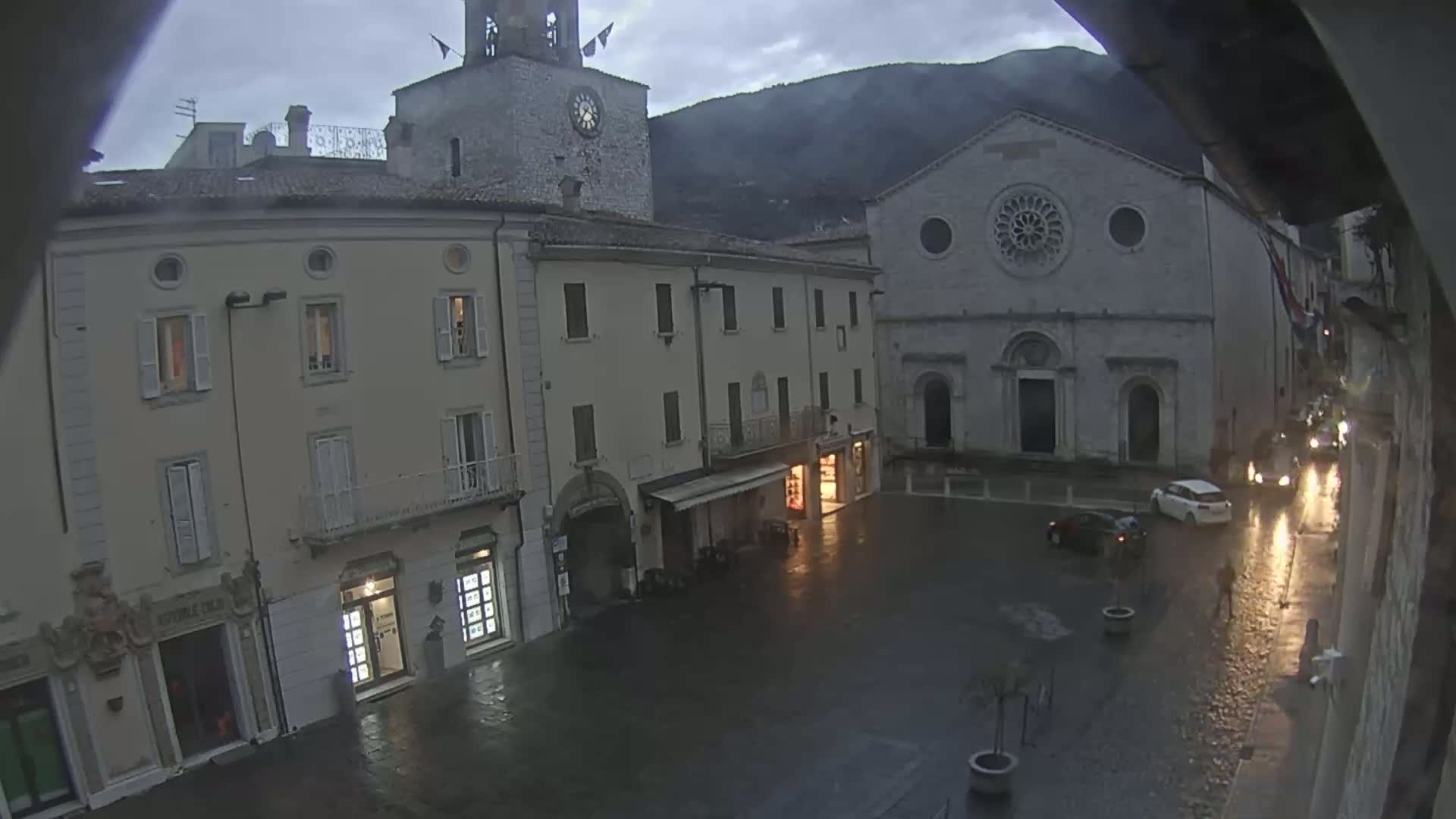 A rain-slicked town square is seen at dusk, with several buildings, including a clock tower and a large church, under a cloudy sky and mountains in the distance.