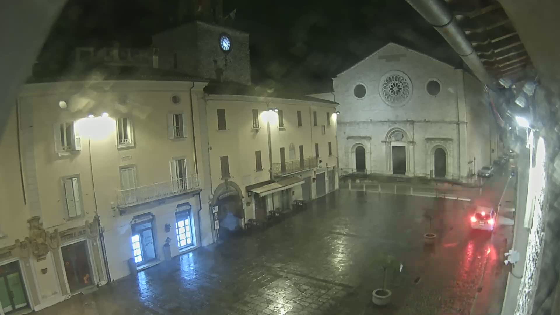 A rain-slicked town square at night, featuring several buildings including a church with a large rose window, is illuminated by streetlights.
