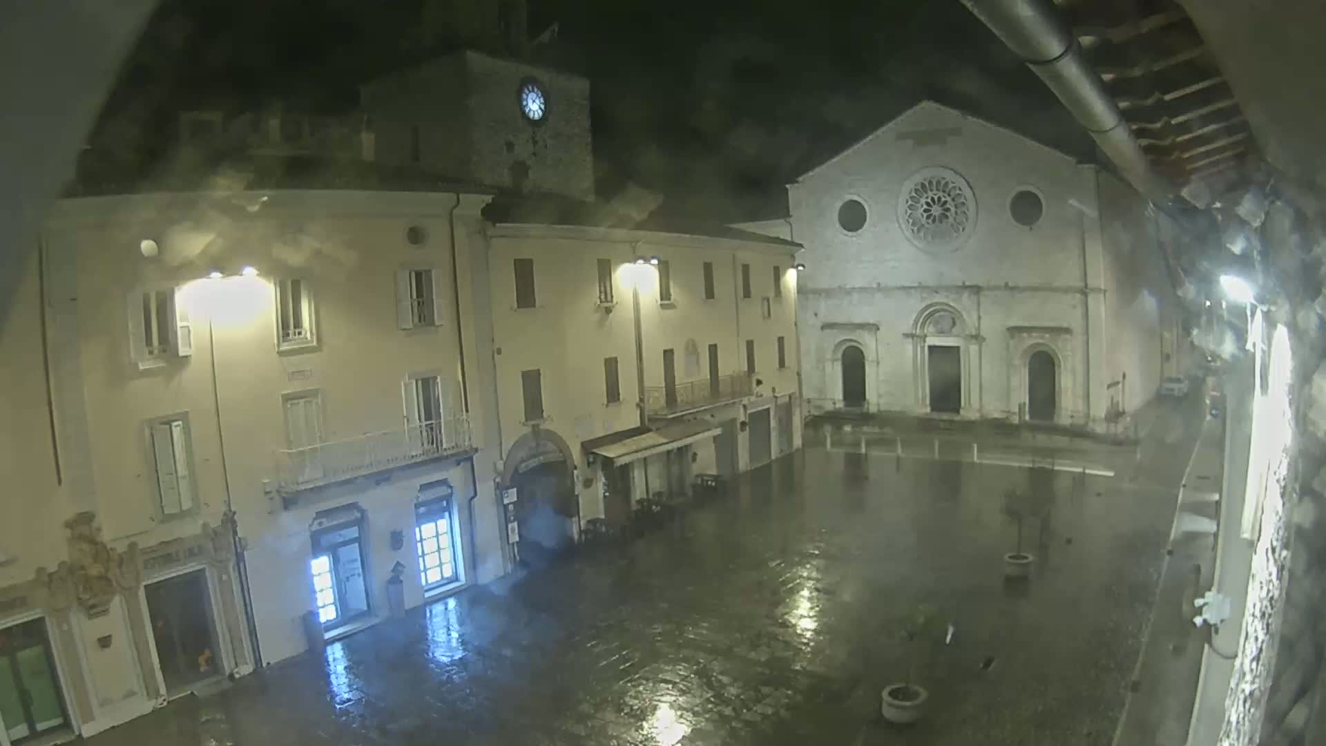A rain-slicked town square at night is illuminated by streetlights, showing several buildings including a church with a large rose window.