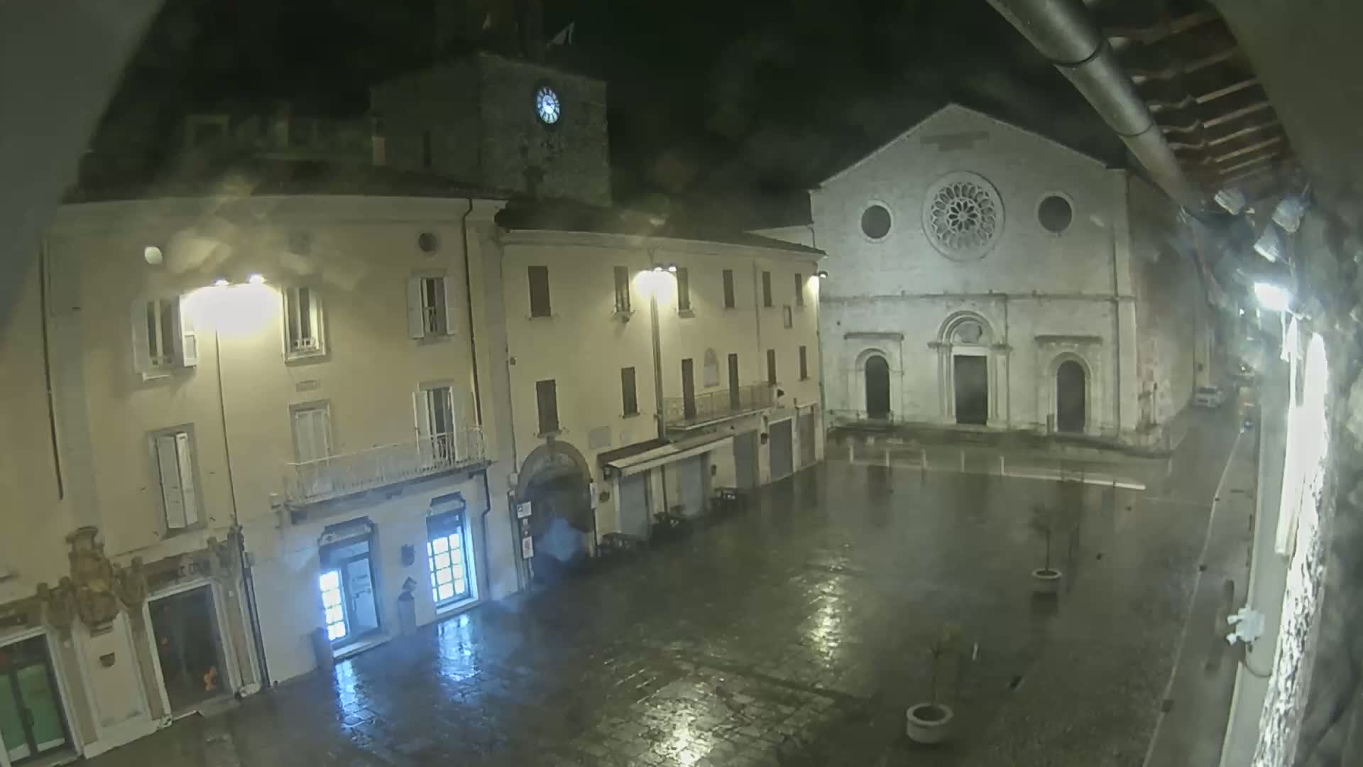 A rain-slicked town square at night, featuring buildings with illuminated windows and a church with a rose window.