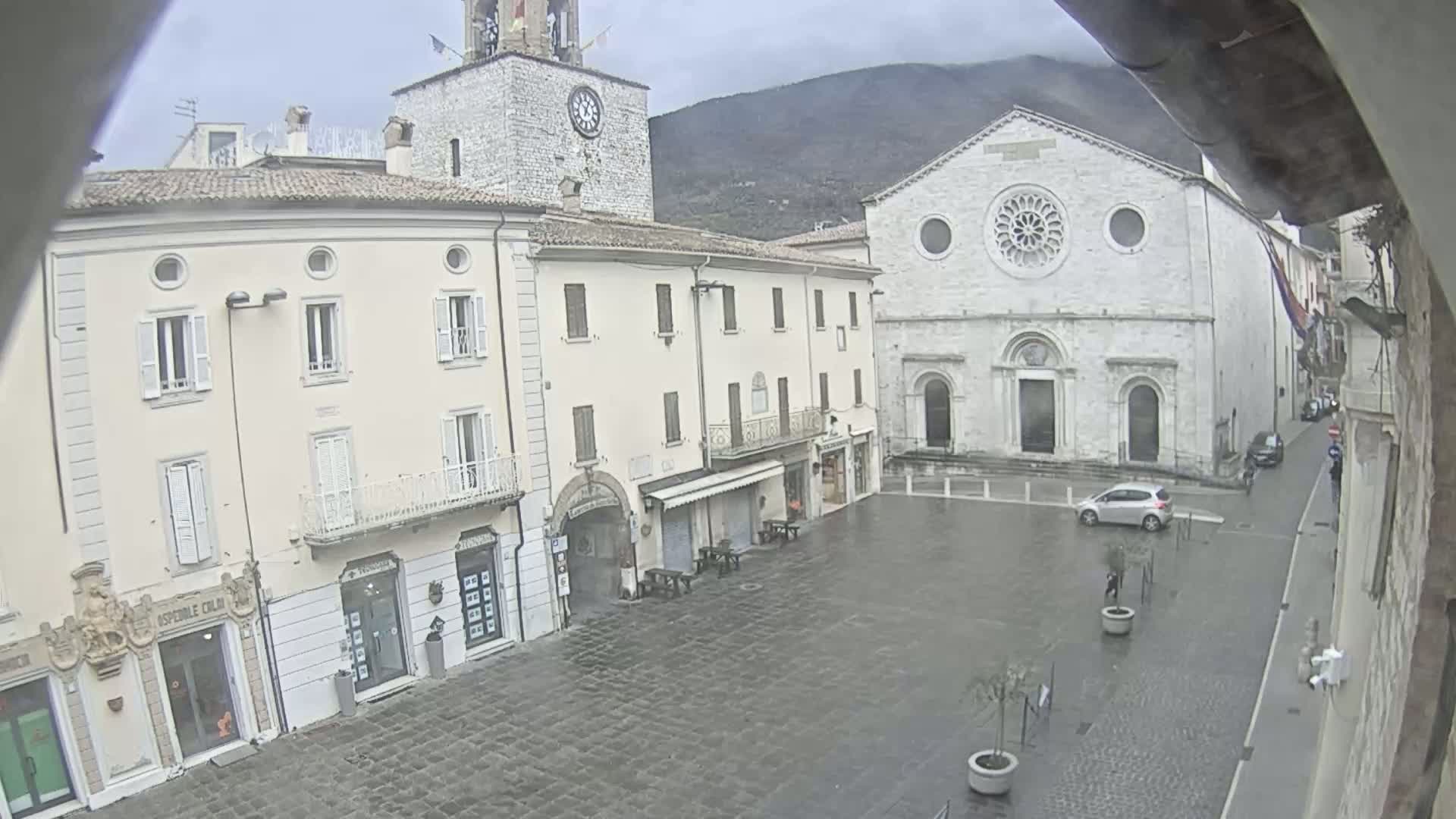 A wet town square is seen with several buildings, including a large church and a clock tower, under an overcast sky.