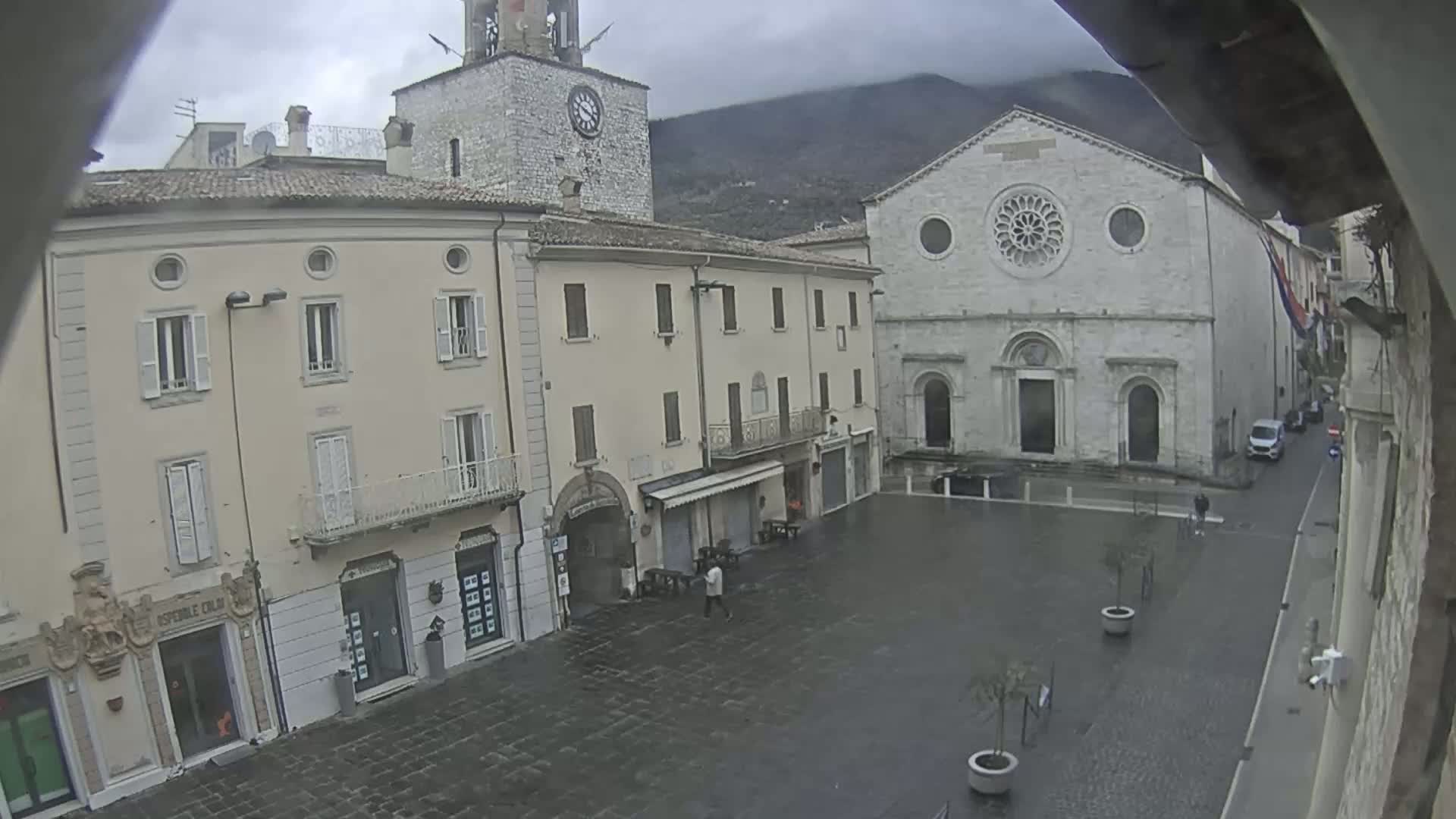 A town square with several buildings, including a church and a clock tower, is shown on an overcast day with wet pavement.