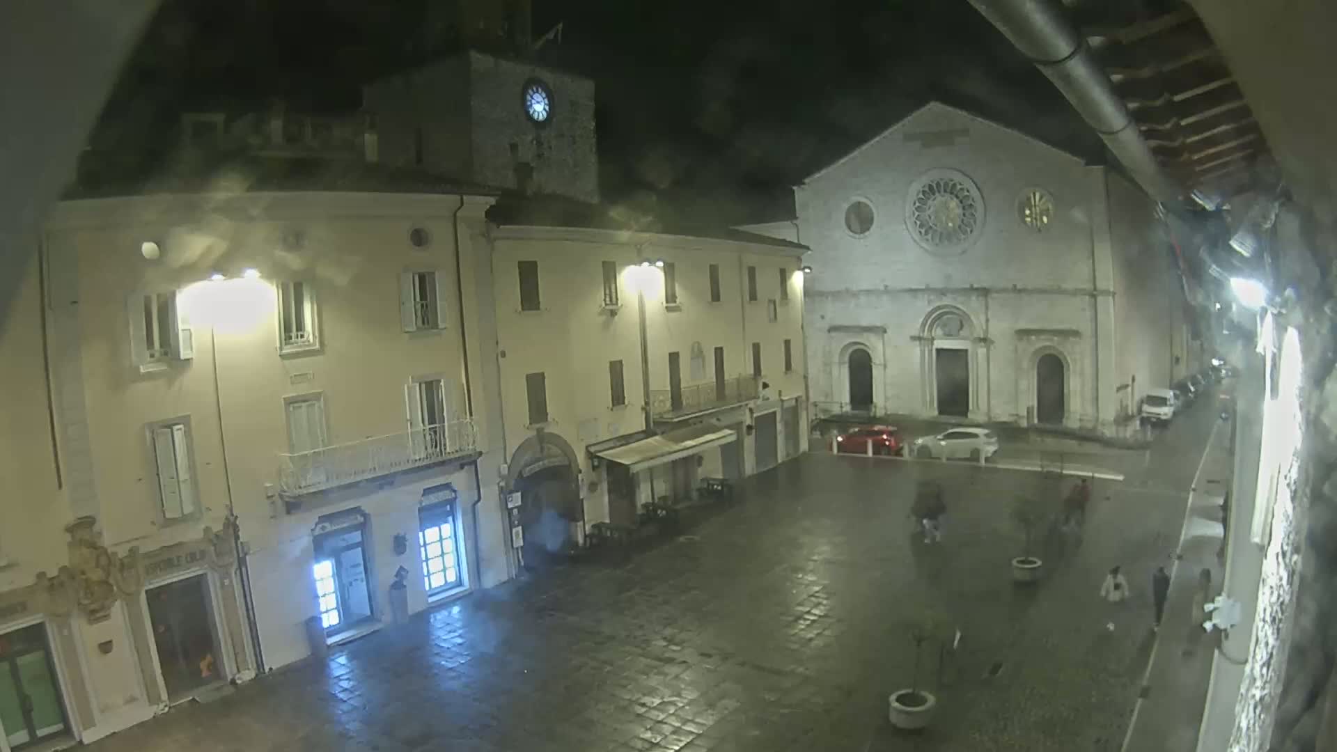 A rain-slicked town square at night is illuminated by streetlights, showing several buildings including a church with a rose window and a clock tower.