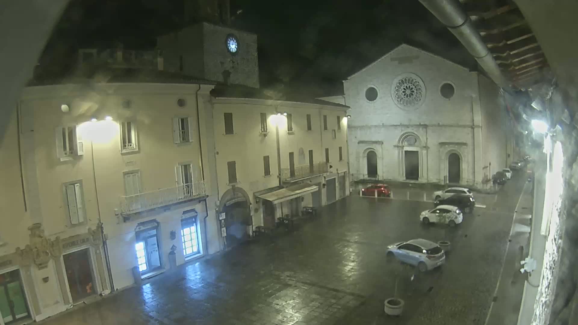 A rain-slicked town square at night is illuminated by streetlights, showing several parked cars and buildings, including a church with a large rose window.