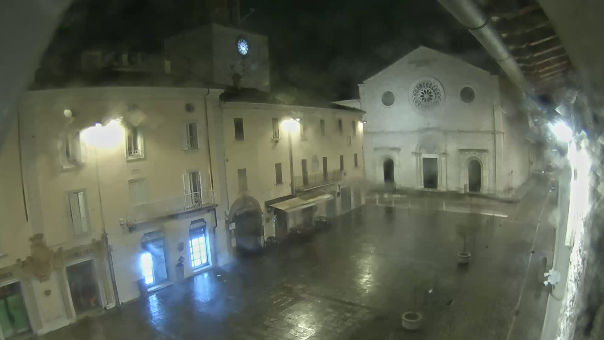 A rain-slicked town square at night is illuminated by streetlights, showing several buildings including a church with a rose window.