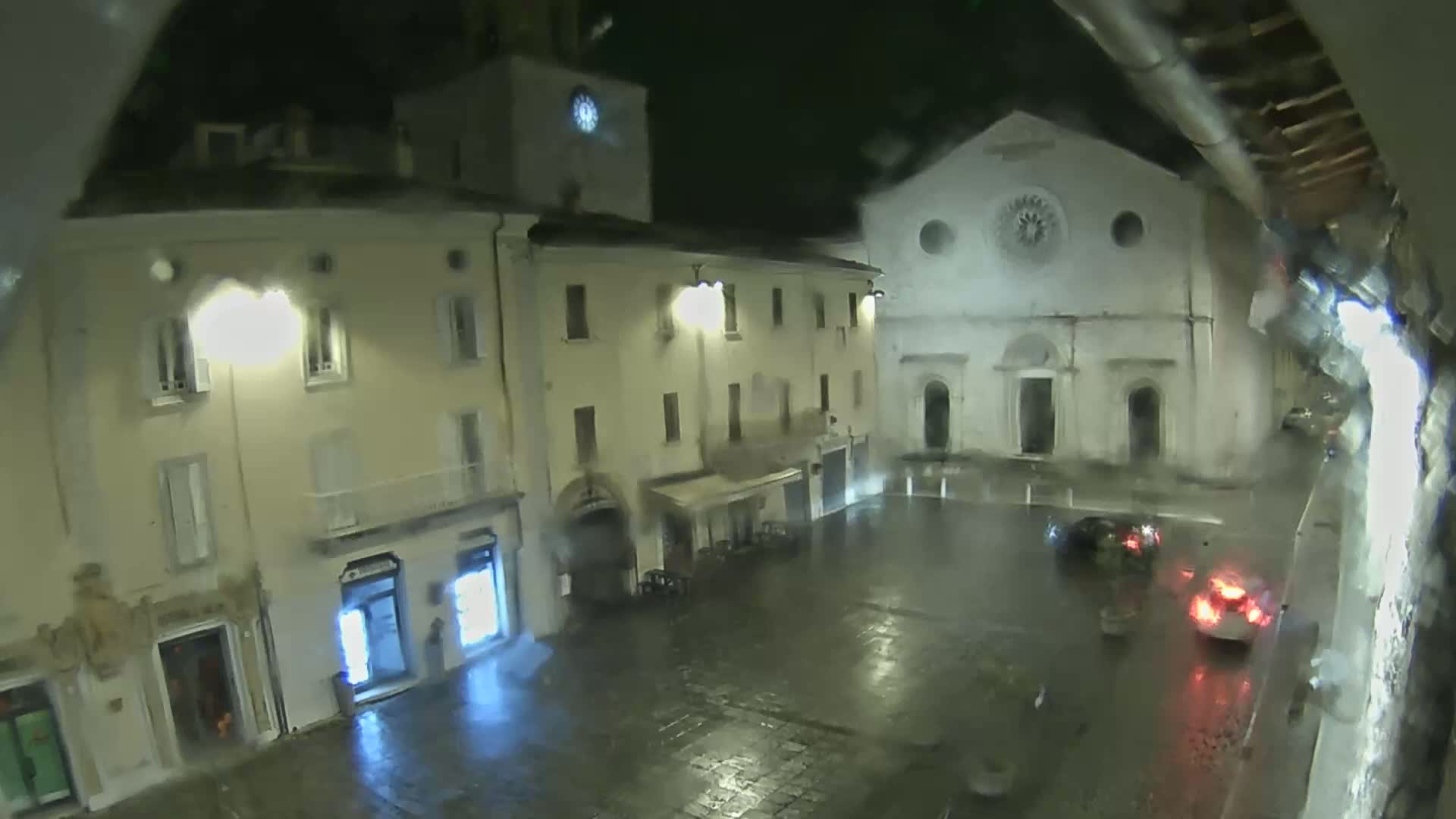 A rain-slicked town square at night is illuminated by streetlights, showing several buildings, including a church with a large circular window, and a few cars parked in the square.