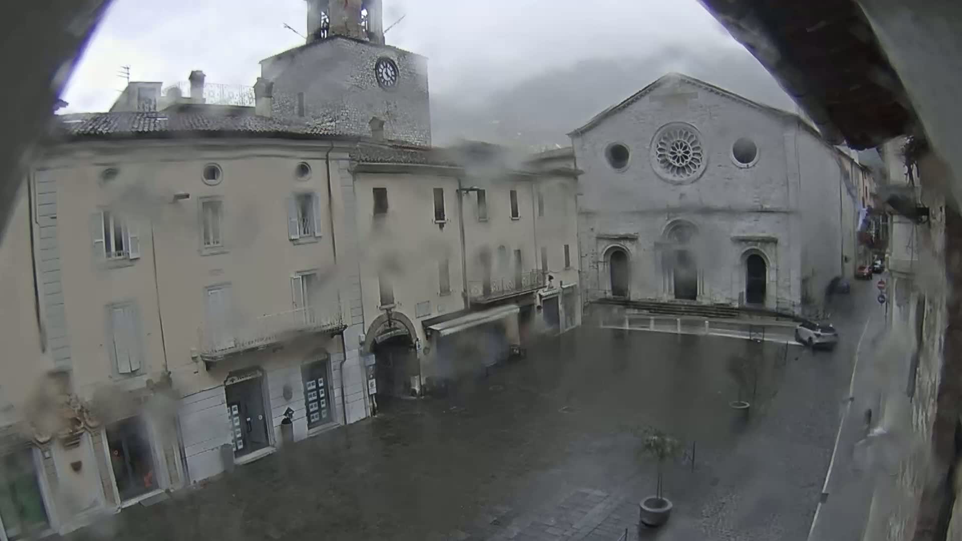 A rain-soaked town square is seen from a high vantage point, featuring buildings with classical architecture, including a clock tower and a church with a rose window.