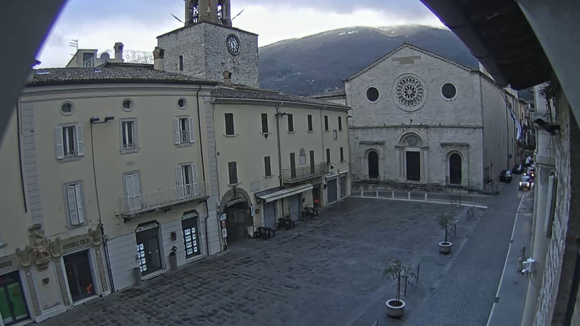 A mostly empty cobblestone town square is flanked by several buildings, including a church with a rose window, under a cloudy sky with mountains in the background.