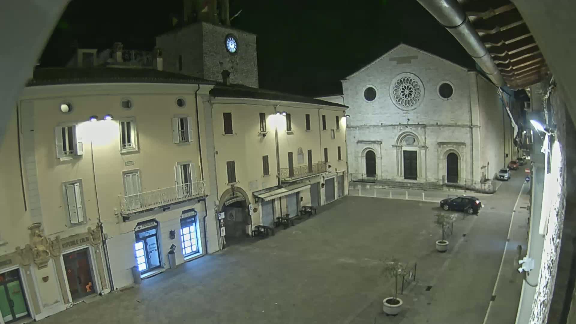 A mostly empty town square at night, illuminated by streetlights, features buildings including a church with a rose window and a clock tower.
