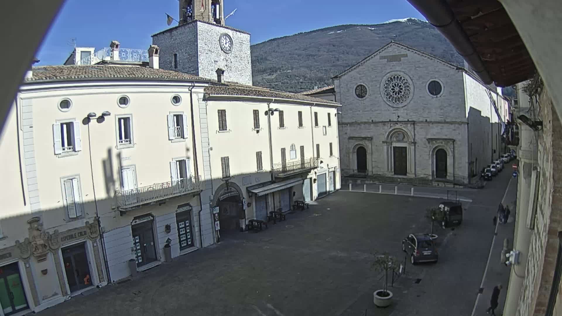 A sunny day reveals a town square with light-colored buildings, including a church with a rose window and a clock tower, under a clear blue sky with mountains in the background.