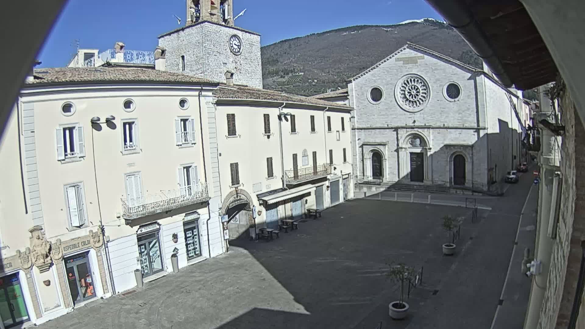 A sunlit town square is shown, featuring several light-colored buildings, including a church with a rose window, and a clock tower, with mountains in the background under a clear blue sky.