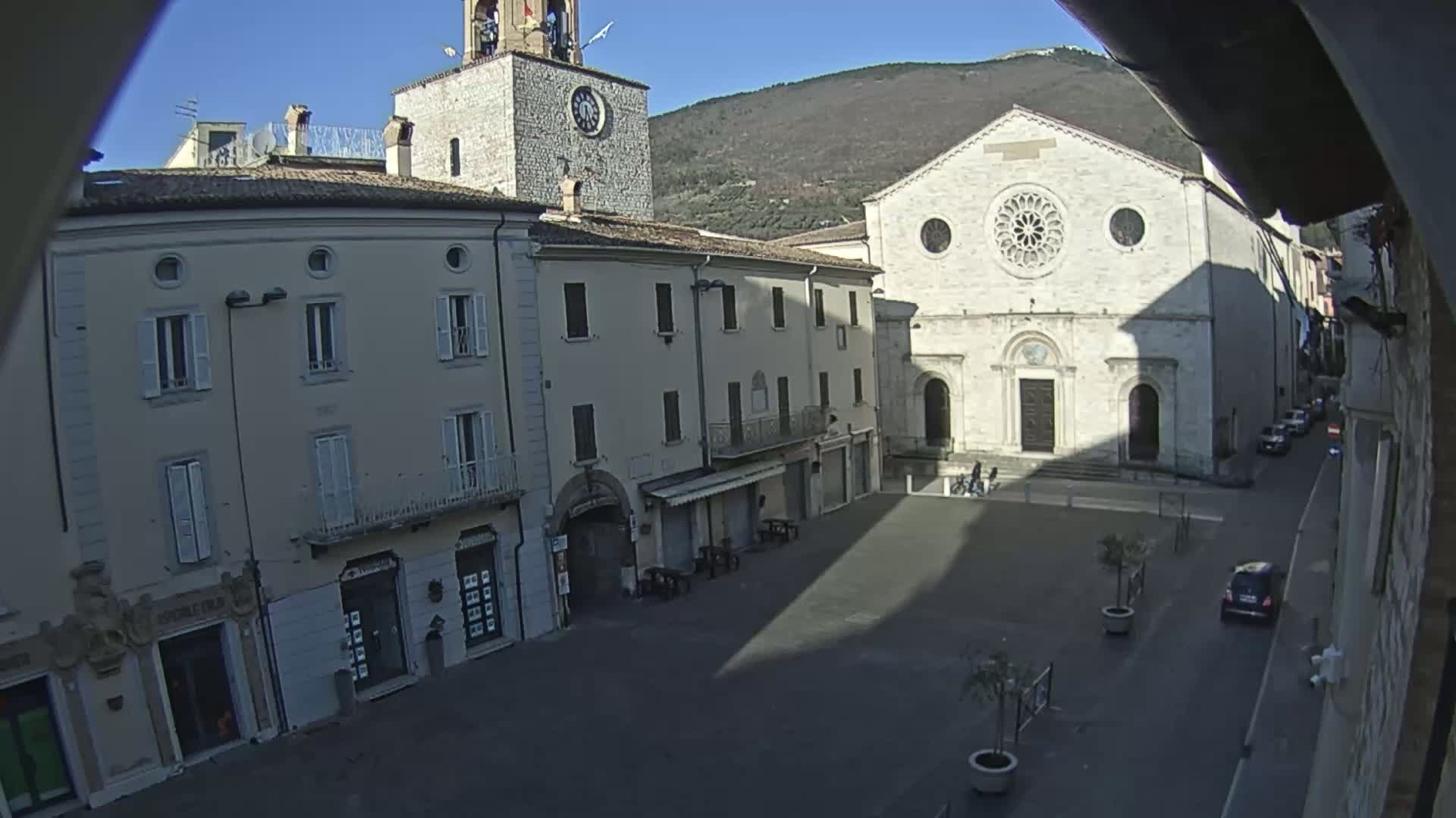 A mostly empty town square is viewed from above on a sunny day, showing several buildings, including a church with a rose window and a clock tower, with a mountain visible in the background.