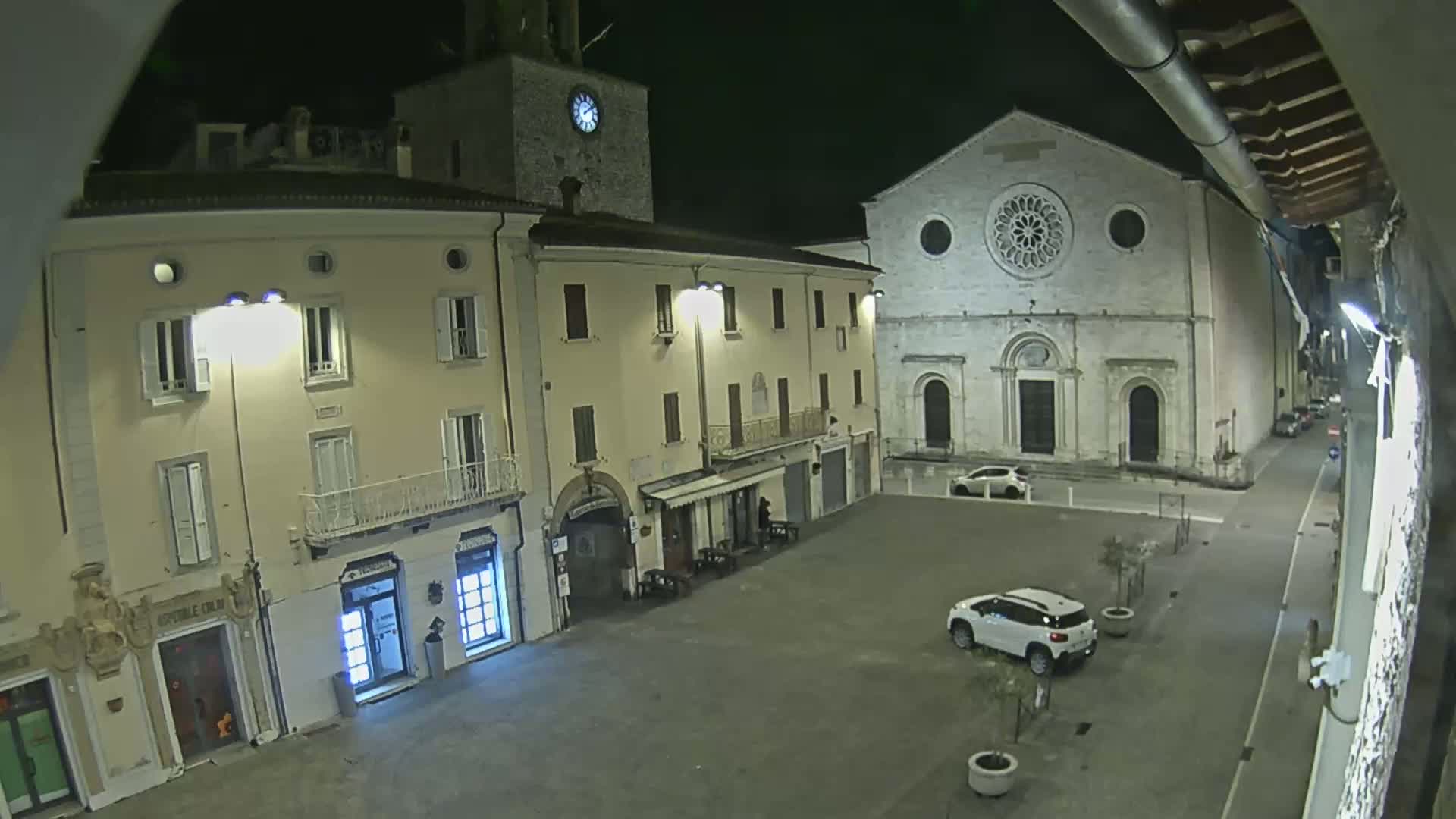 A nighttime view of a town square with buildings, including a church with a rose window, and a few parked cars.