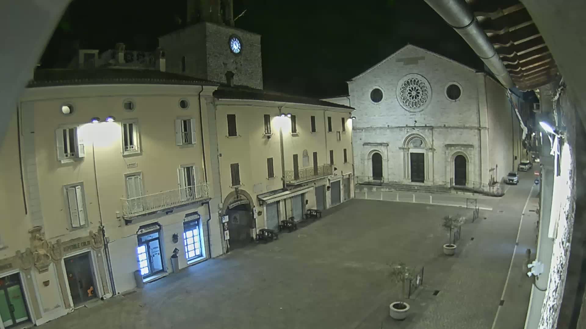 A nighttime view of an empty town square, featuring several buildings including a church with a large rose window, and a clock tower.