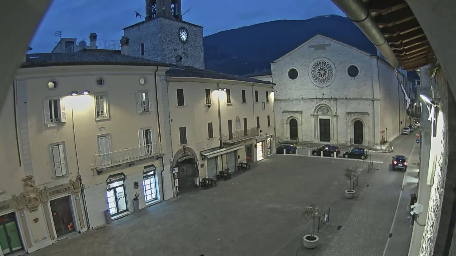 A town square at night, under a twilight sky, is illuminated by streetlights and features a church with a large rose window and a clock tower.