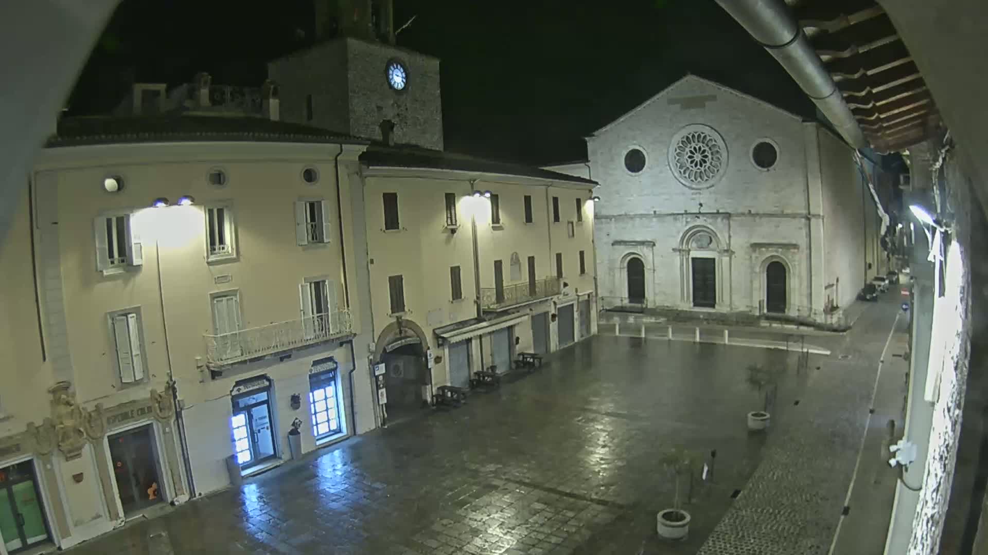 A wet, empty town square at night is bordered by buildings, including a church with a large rose window and a clock tower.