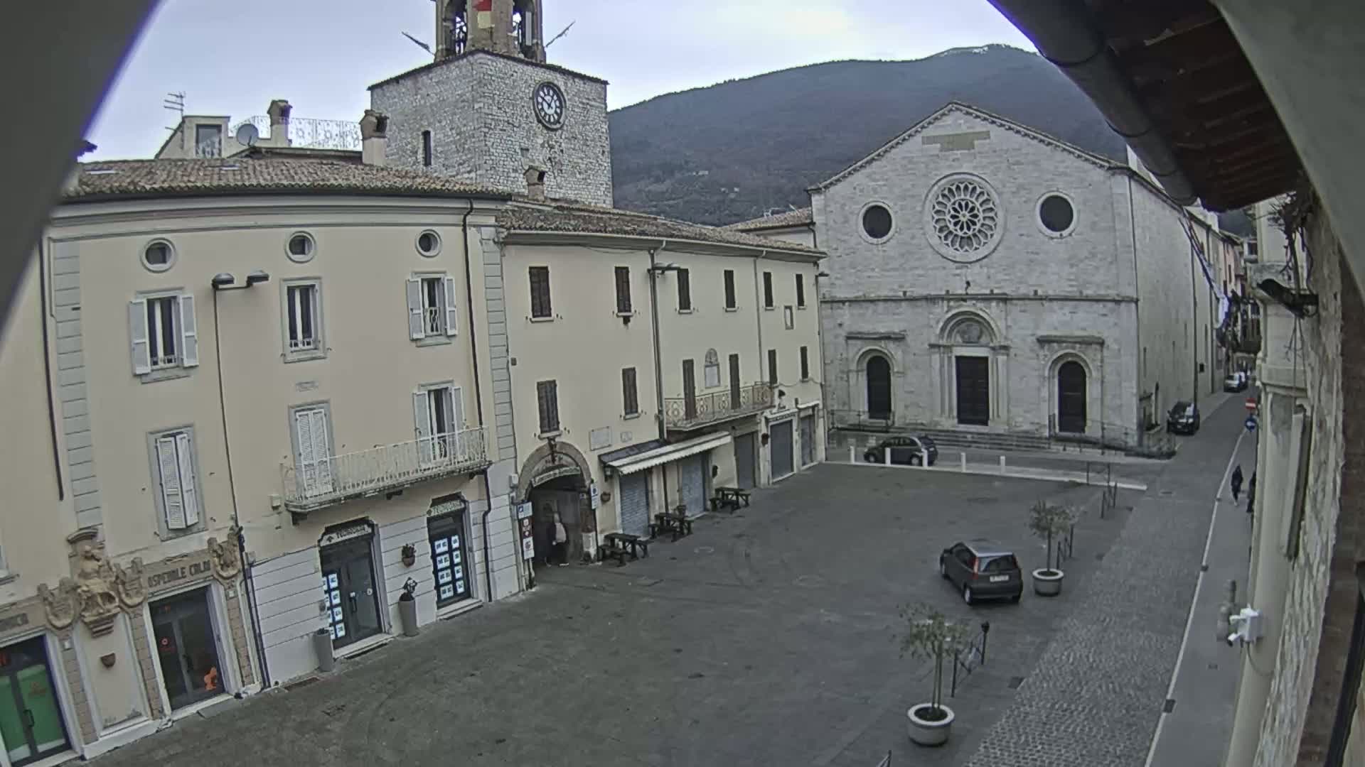 A mostly empty town square is situated between several buildings, including a clock tower and a large stone church, under an overcast sky.