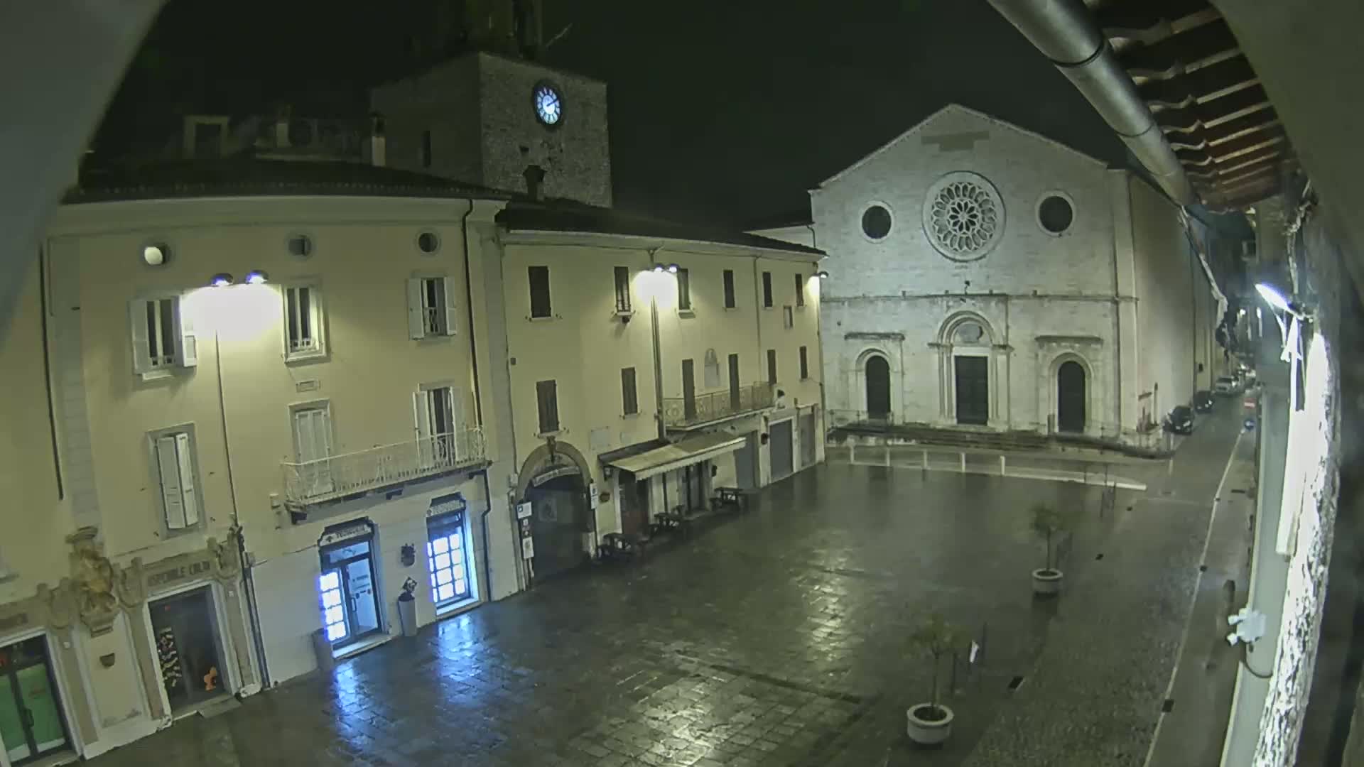 A rain-slicked town square at night, illuminated by streetlights, features buildings including a clock tower and a large church with a rose window.