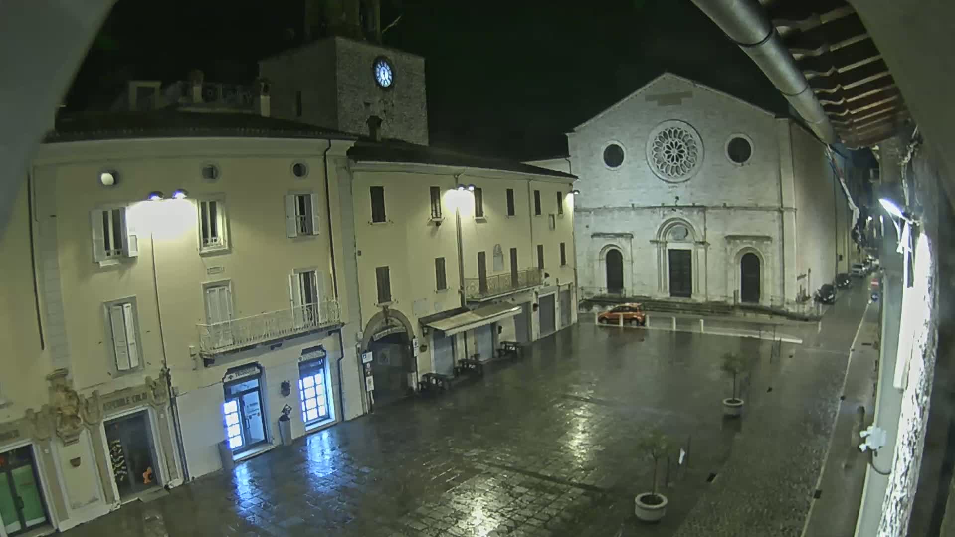 A rain-slicked town square at night, featuring a clock tower, a large church, and several buildings, all illuminated by streetlights.