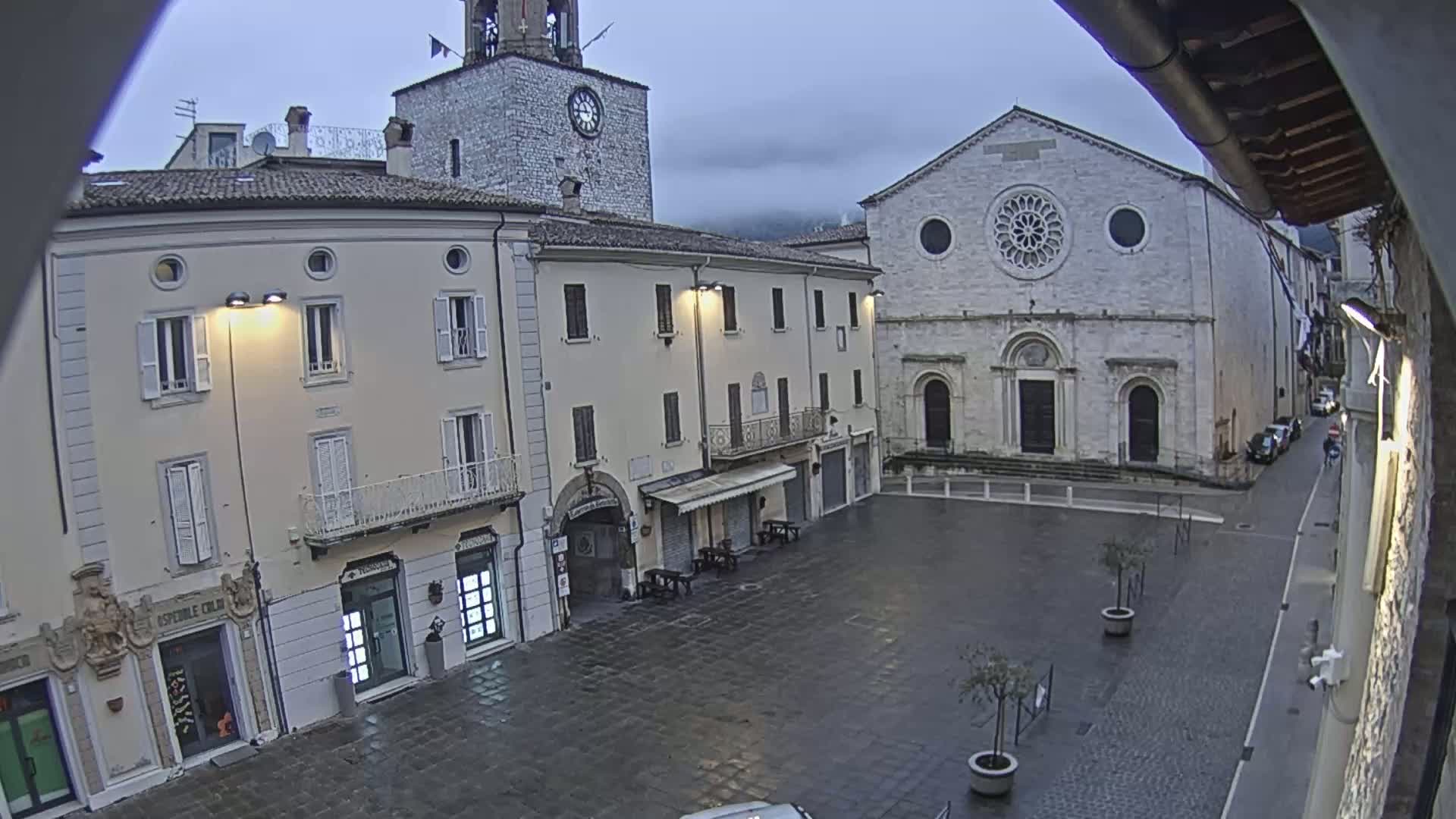 A mostly empty town square is seen on an overcast day, with stone buildings, including a church with a rose window, and a clock tower visible.