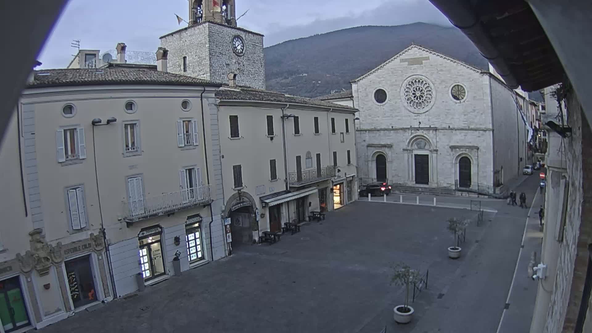 An overcast day reveals a town square with light-colored buildings, including a church with a rose window and a clock tower, backed by a mountain range.