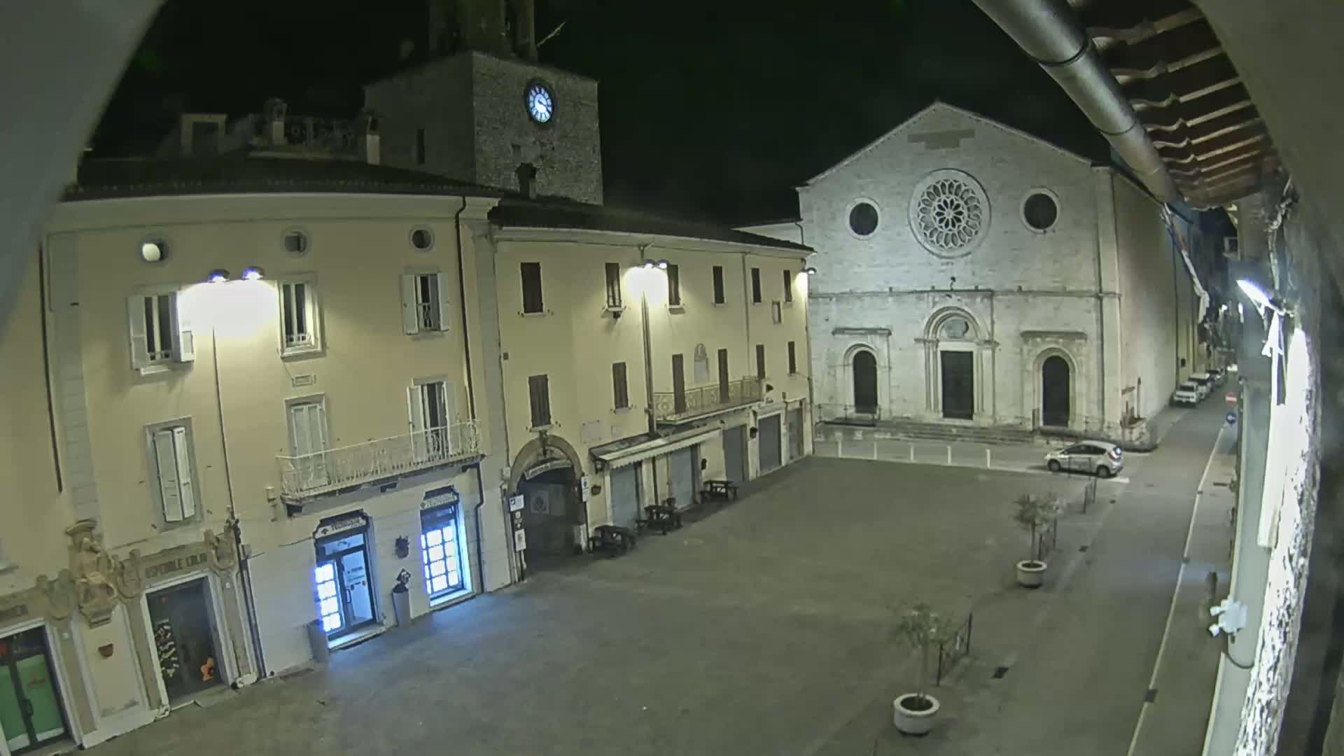 A nighttime view of a town square, showing several buildings including a church with a rose window and a clock tower, under clear skies.