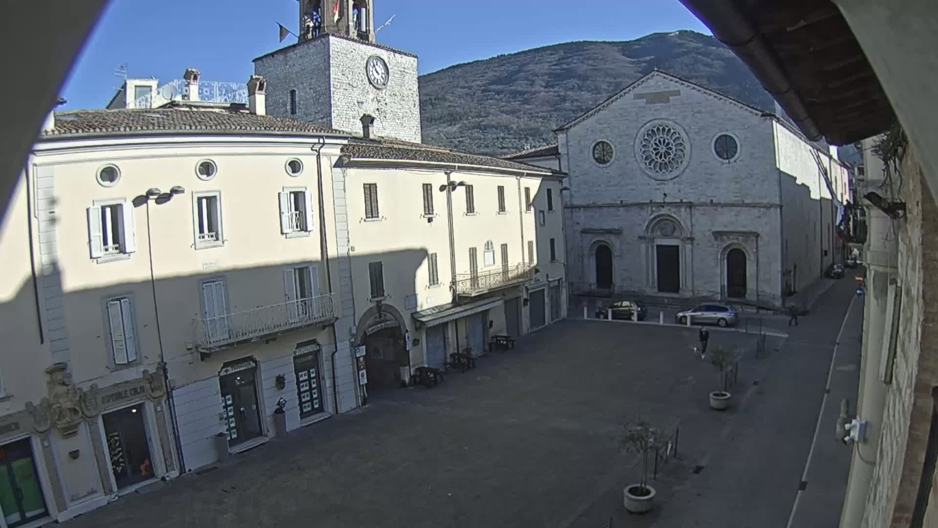 A town square with several light-colored buildings, including a clock tower and a church, is seen on a sunny day with mountains in the background.