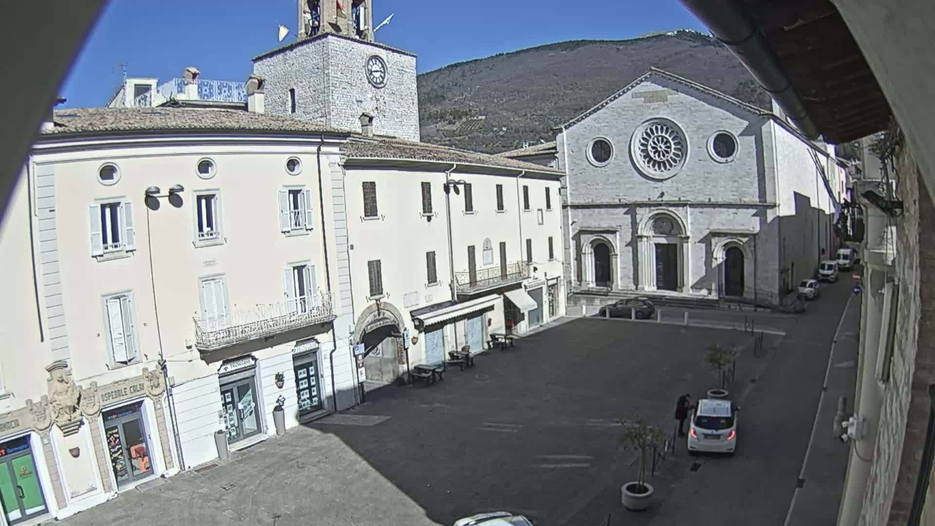 A sunny day in a town square shows several buildings, including a church with a large rose window, and a few cars parked near the edge of the square.