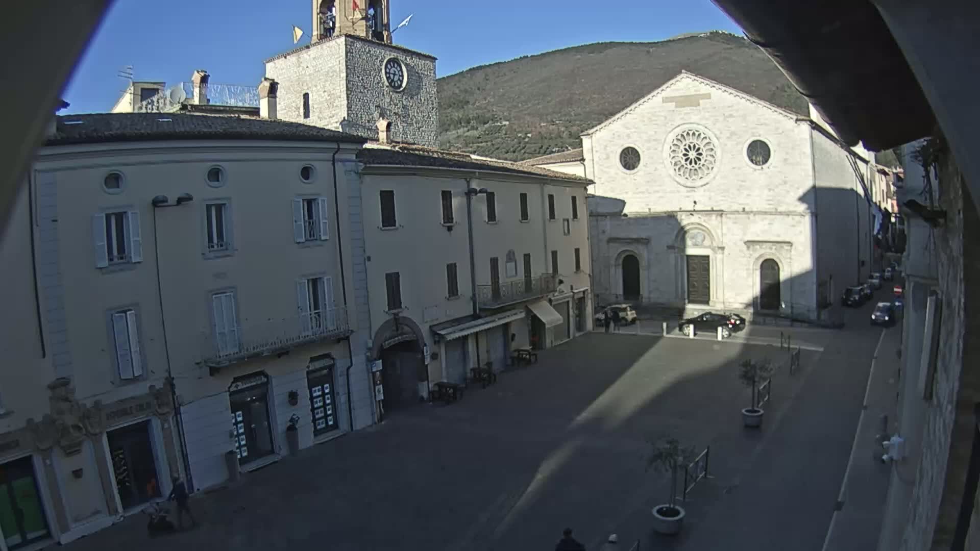 A sunny, high-angle view of a town square featuring several multi-story buildings, one of which has a large rose window, with a mountain visible in the background.