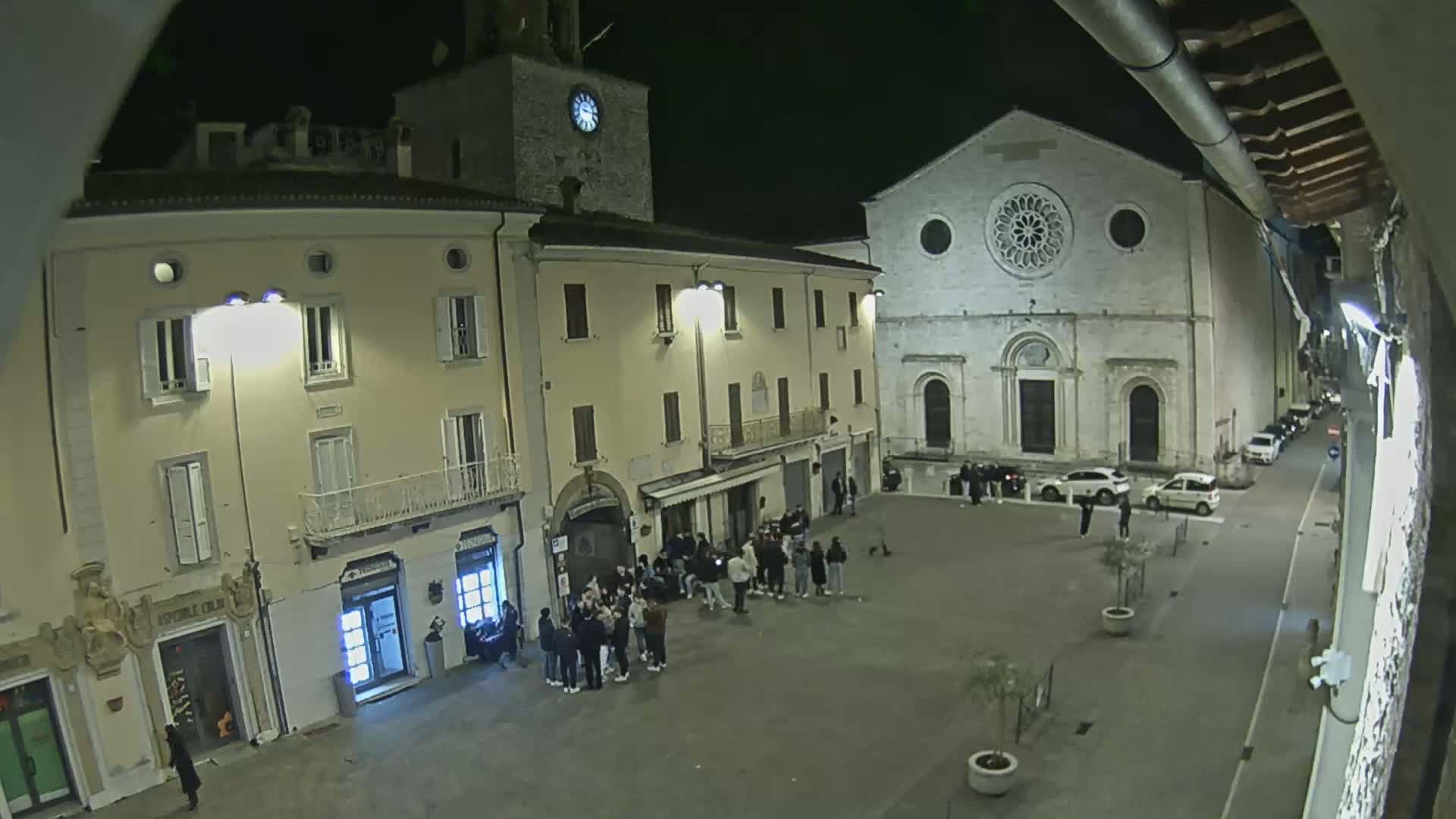 A nighttime view of a town square with several buildings, including a church with a rose window, and a group of people gathered in the center.