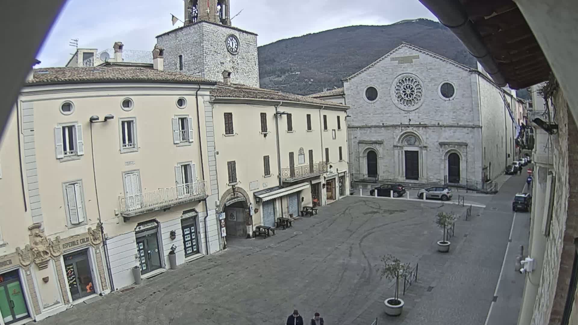 A mostly empty town square is shown on an overcast day, with several buildings surrounding it, including a clock tower and a large church.