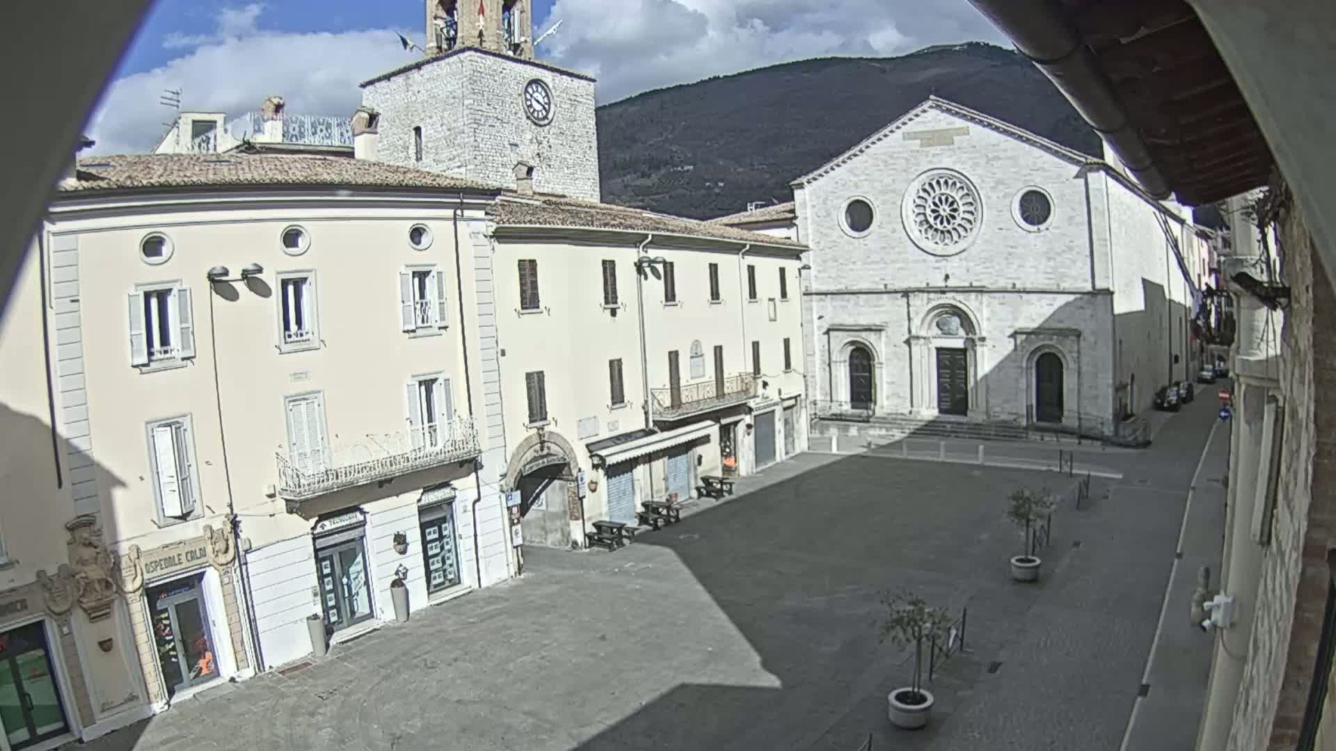 A mostly empty town square on a partly sunny day is surrounded by light-colored buildings, including a church with a large rose window, and a clock tower.