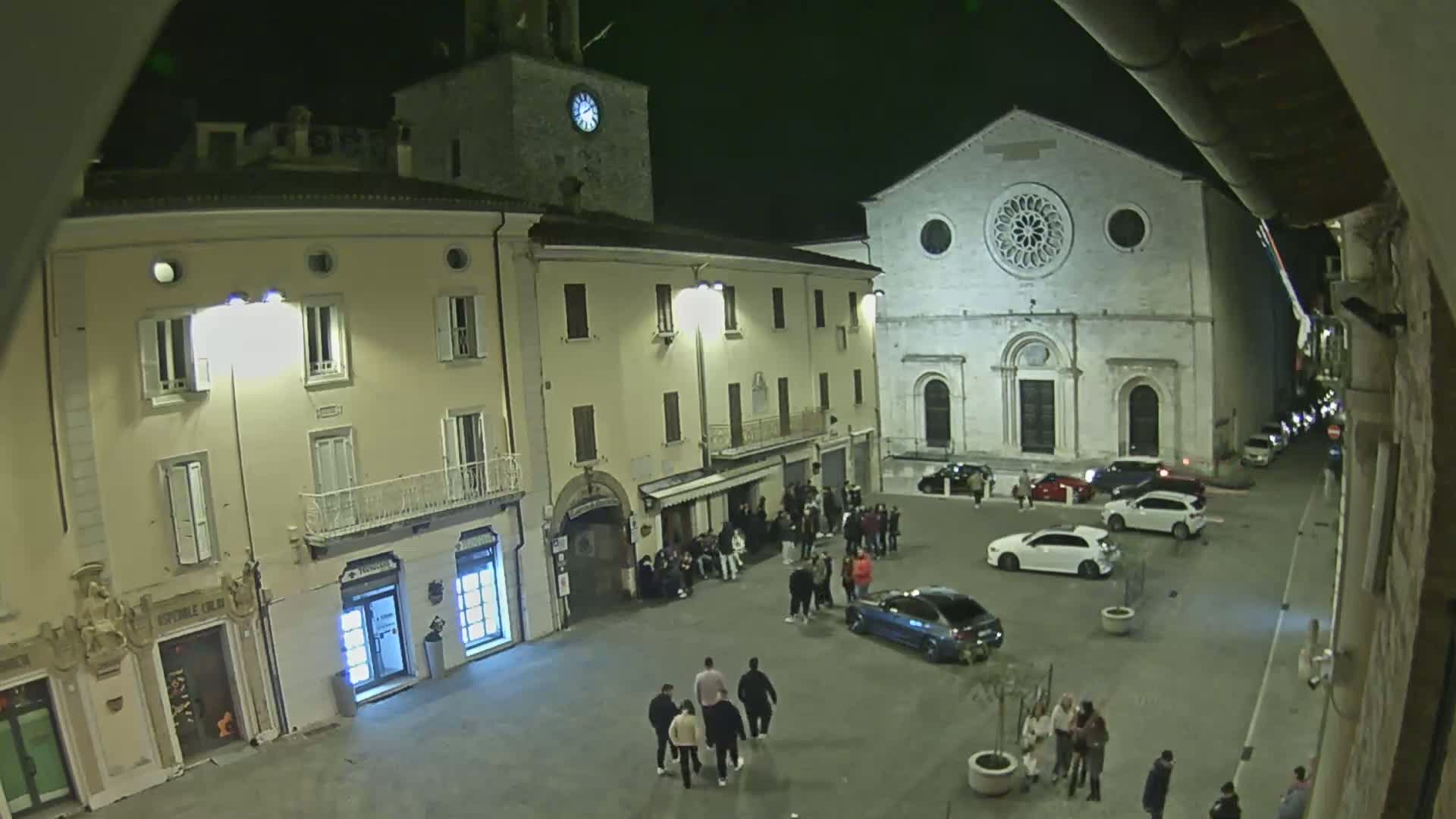 A nighttime, dimly lit town square features several buildings, including a church with a large rose window, and groups of people standing around.