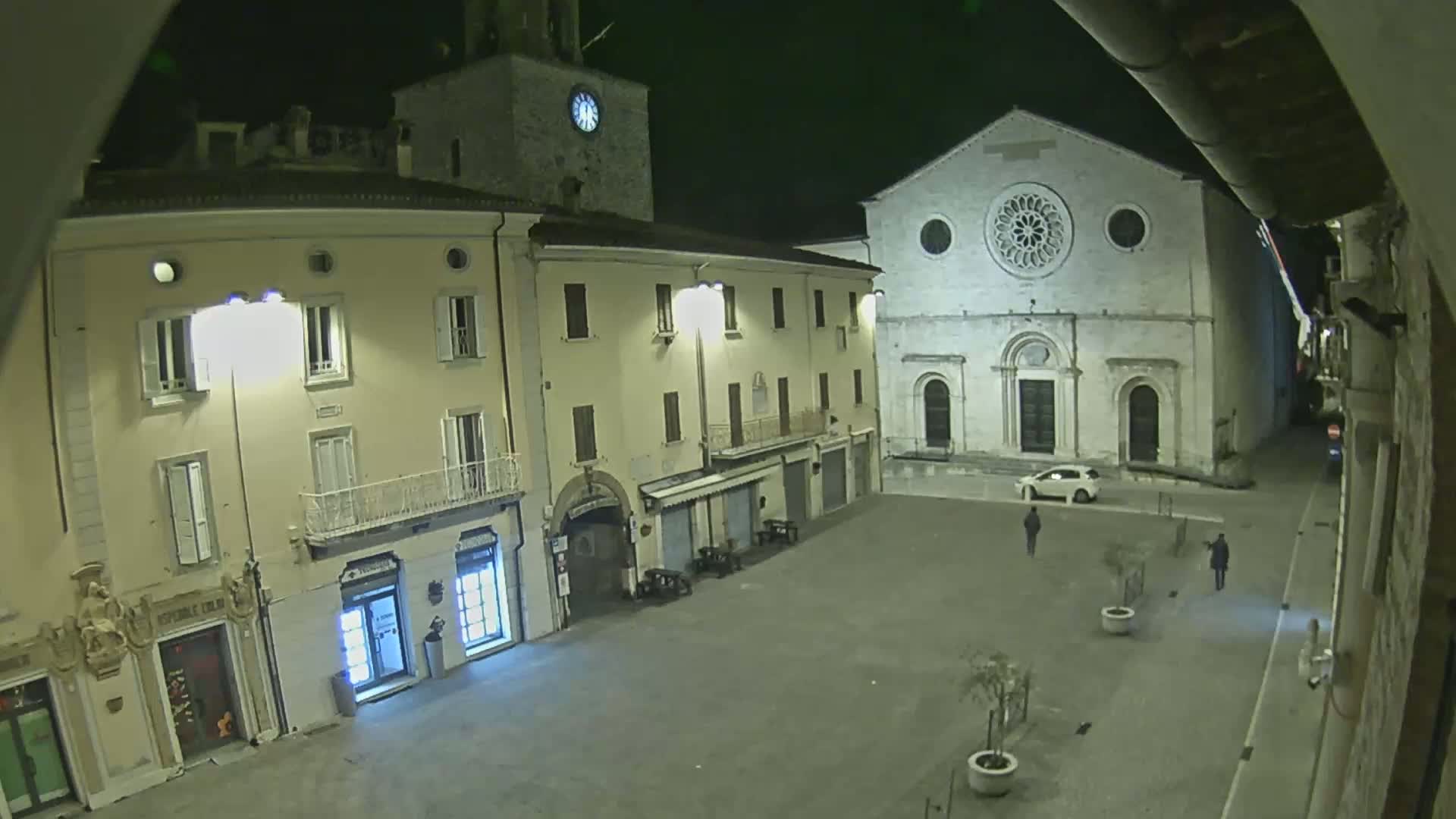 A mostly empty, paved town square at night is illuminated by streetlights, showing several buildings including a church with a rose window, and a few pedestrians.