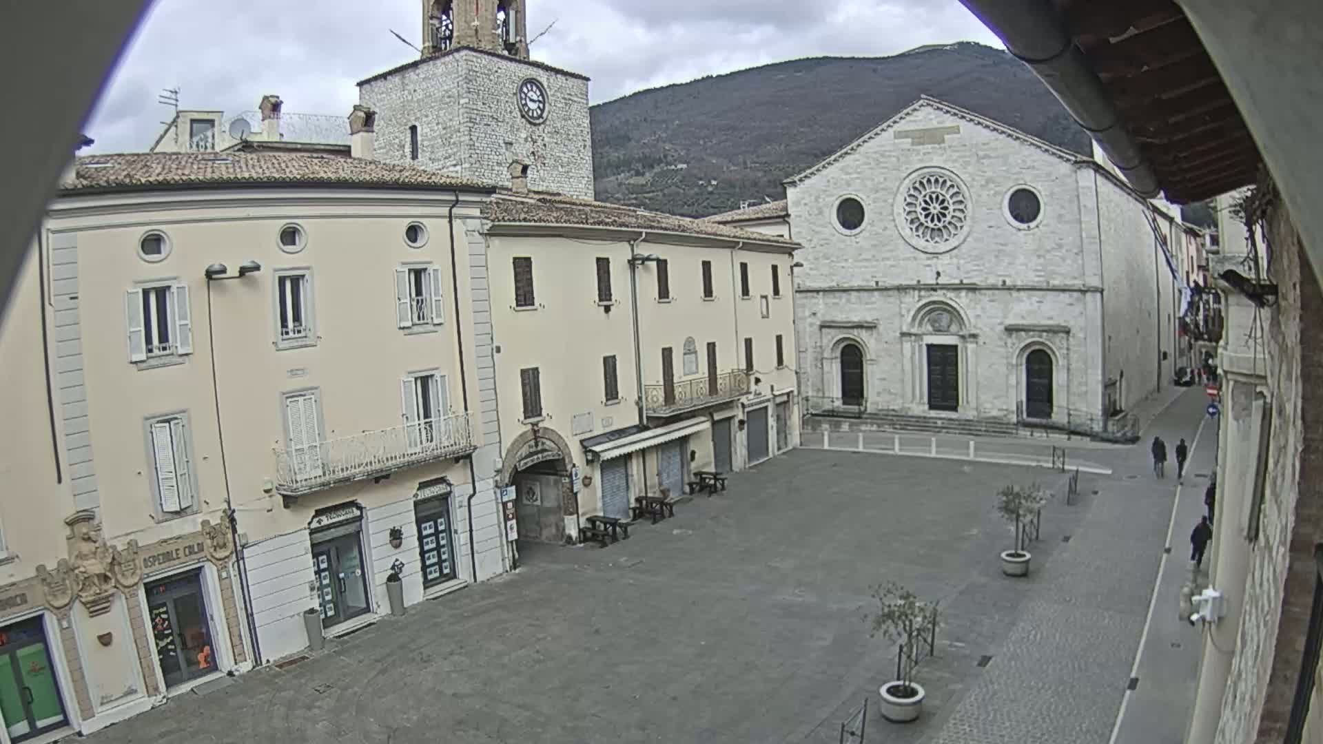 A mostly empty town square is seen on an overcast day, with several buildings surrounding it, including a large stone church and a clock tower.