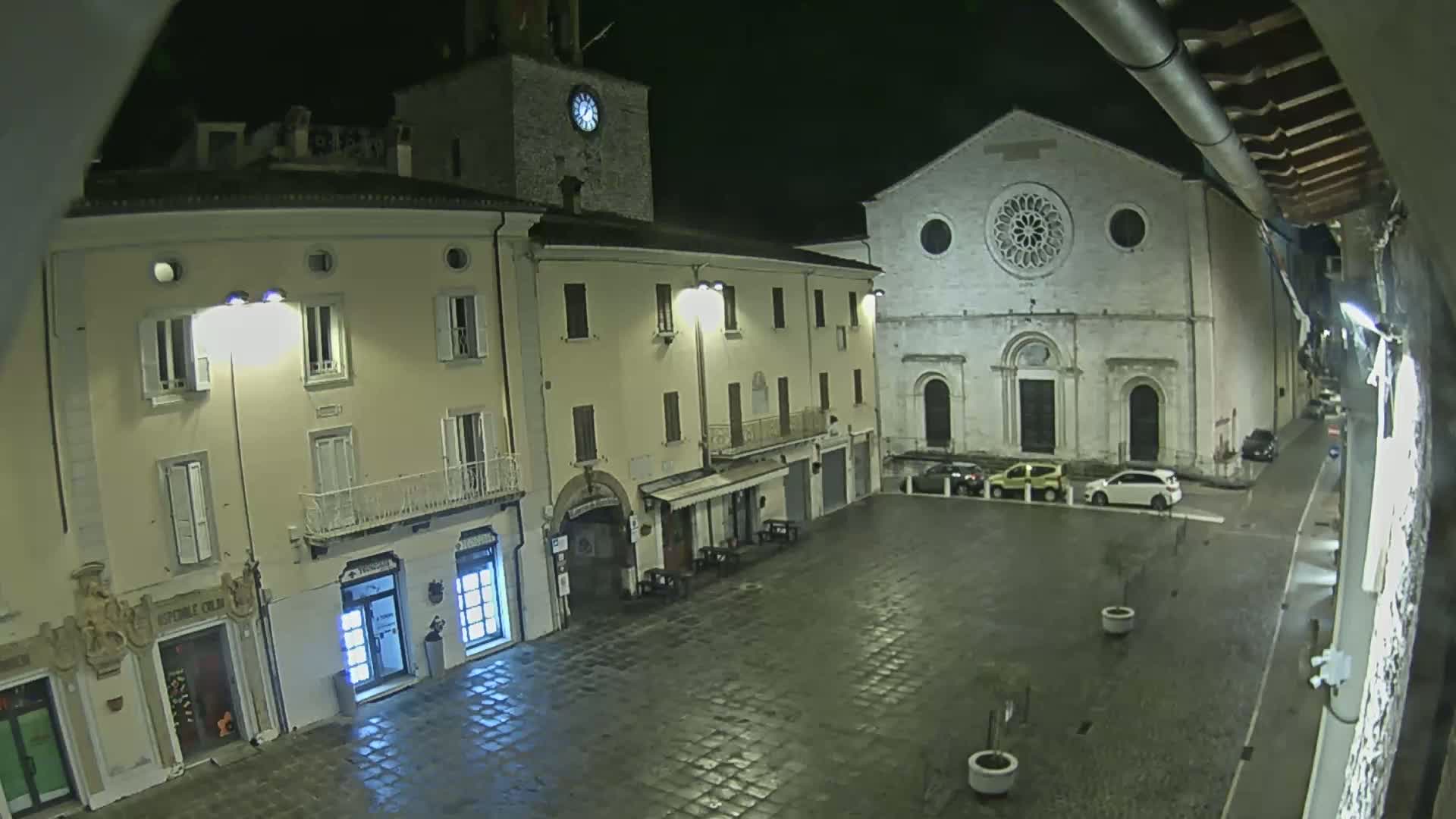 A nighttime view of a town square with several buildings, including a church with a large rose window, lit by streetlights on a wet, paved surface.