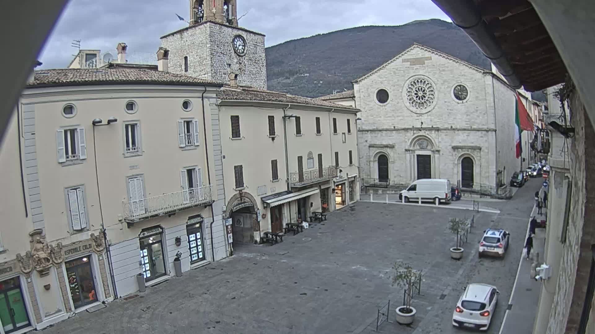 A town square with several buildings, including a church and a clock tower, is seen on an overcast day, with a few cars parked in the square and a mountain visible in the background.