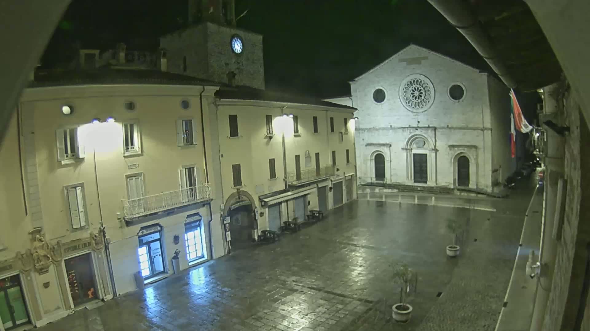 A rain-slicked town square at night, illuminated by streetlights, features several buildings, including a church with a rose window and a clock tower.