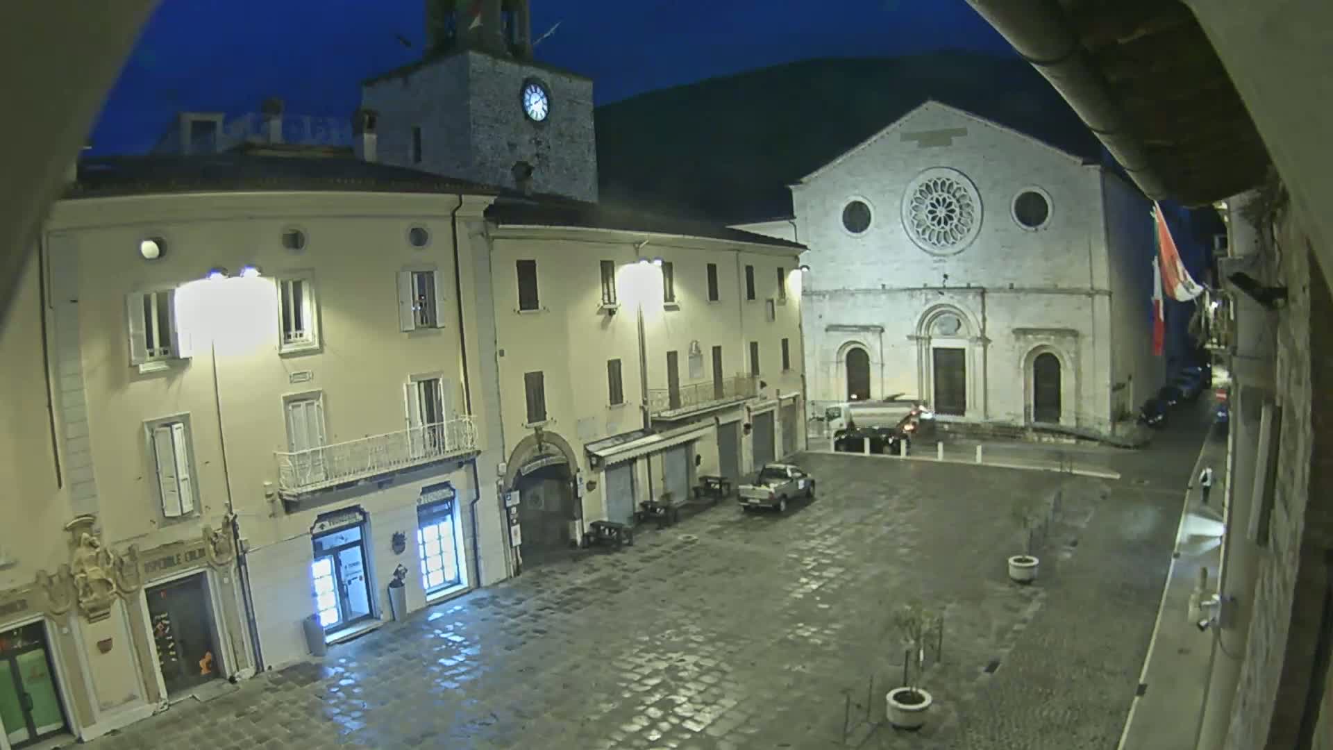 A nighttime view of a town square, illuminated by streetlights, features a large church, several buildings, and a few parked vehicles under a dark sky.