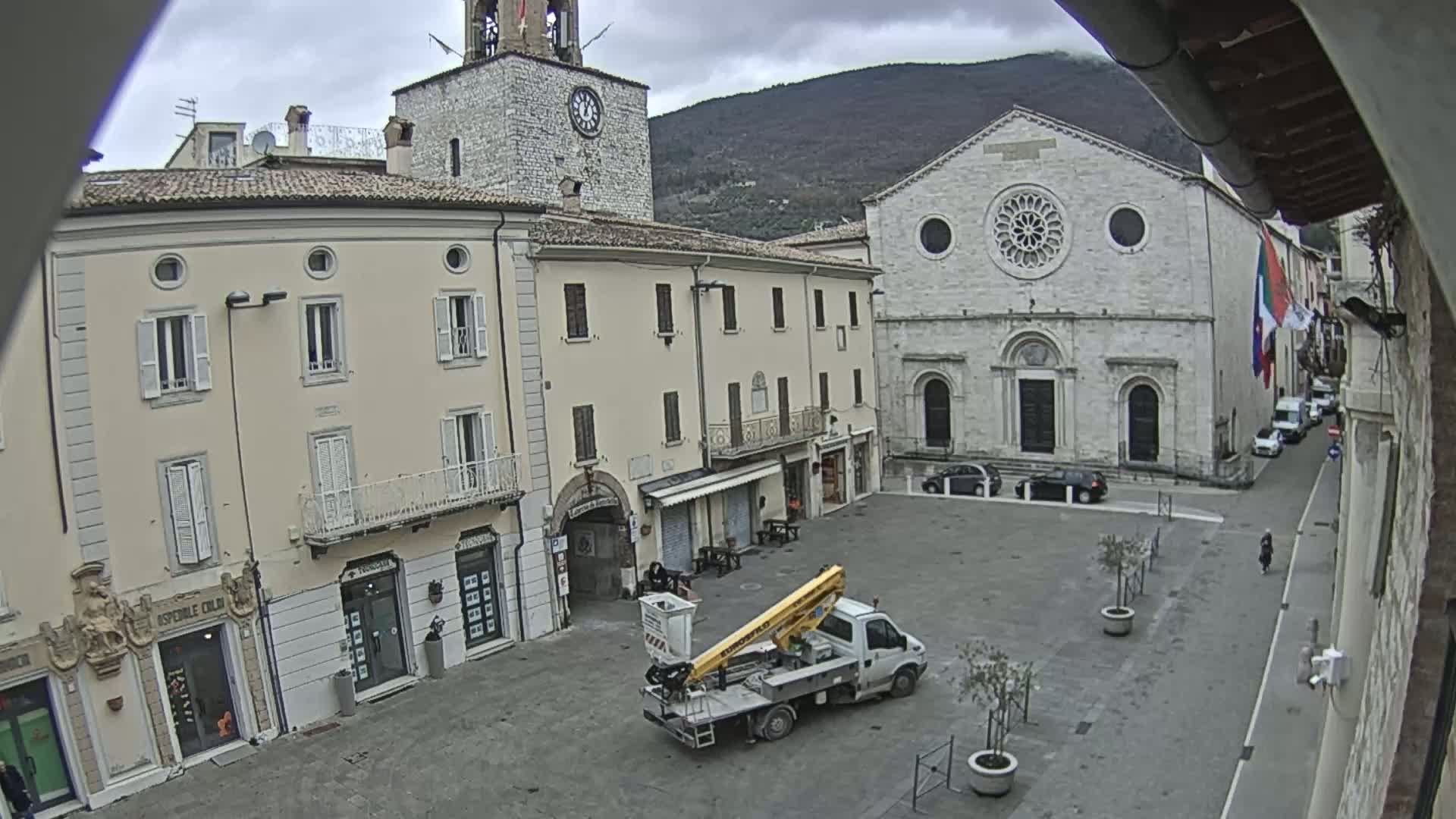 A town square is seen on an overcast day, with several buildings including a church and a clock tower surrounding it, and a small bucket truck parked in the center.