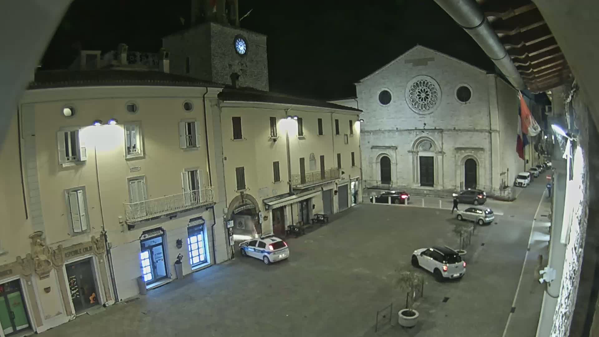 A nighttime, high-angle view of a town square with several buildings, including a church with a rose window, and a few parked cars.
