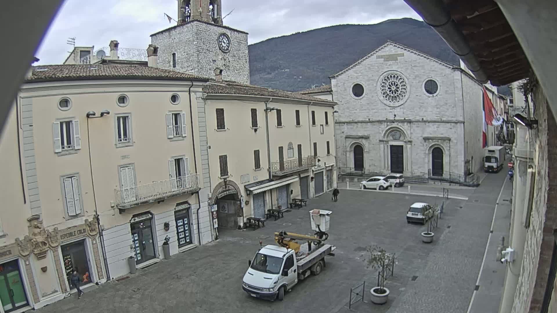 A mostly empty town square under an overcast sky features several light-colored buildings, including a church with a rose window, a clock tower, and shops, with a few parked cars and a small maintenance truck.