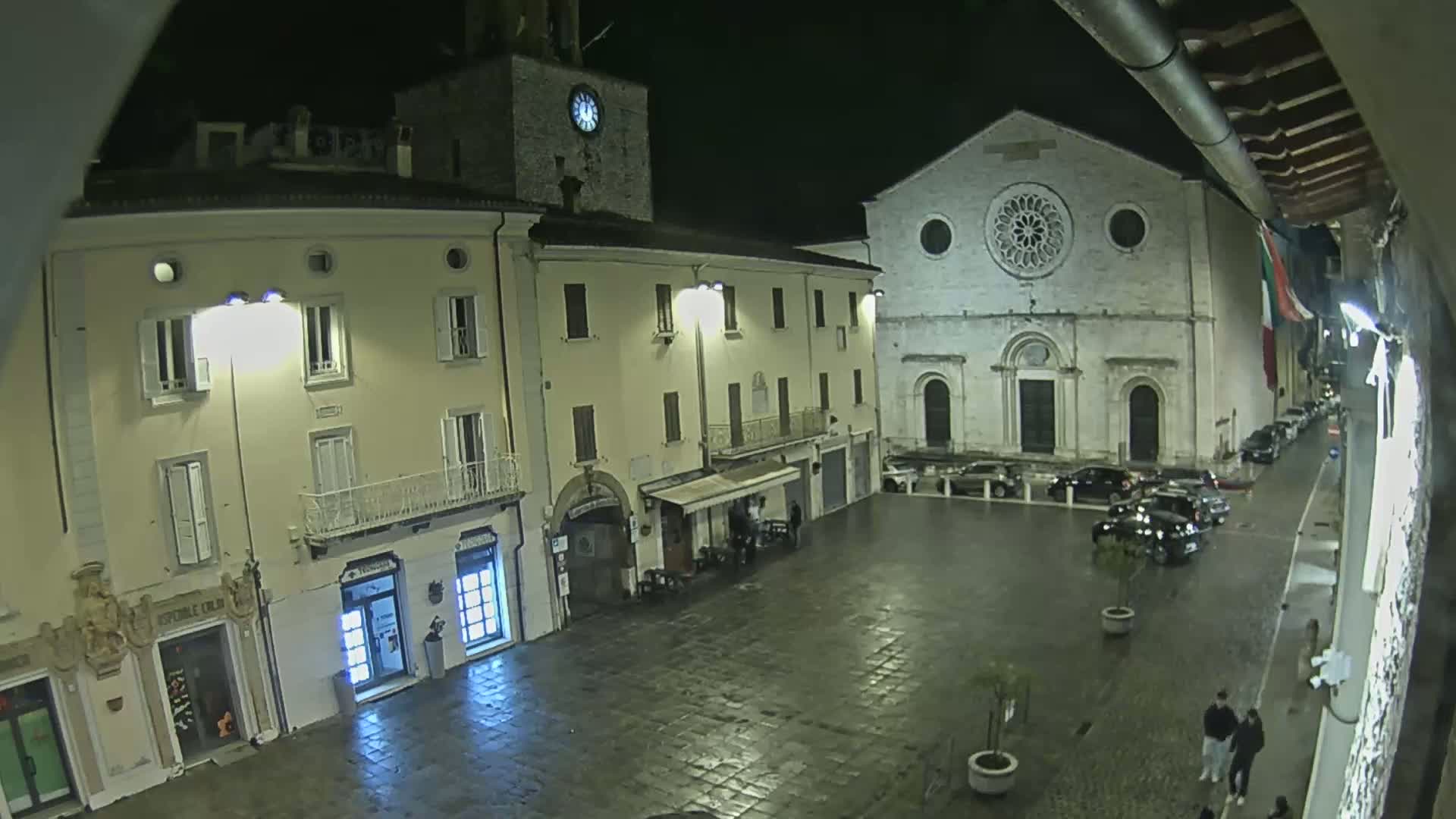A nighttime view of a town square with several buildings, including a church with a large rose window, under wet conditions.