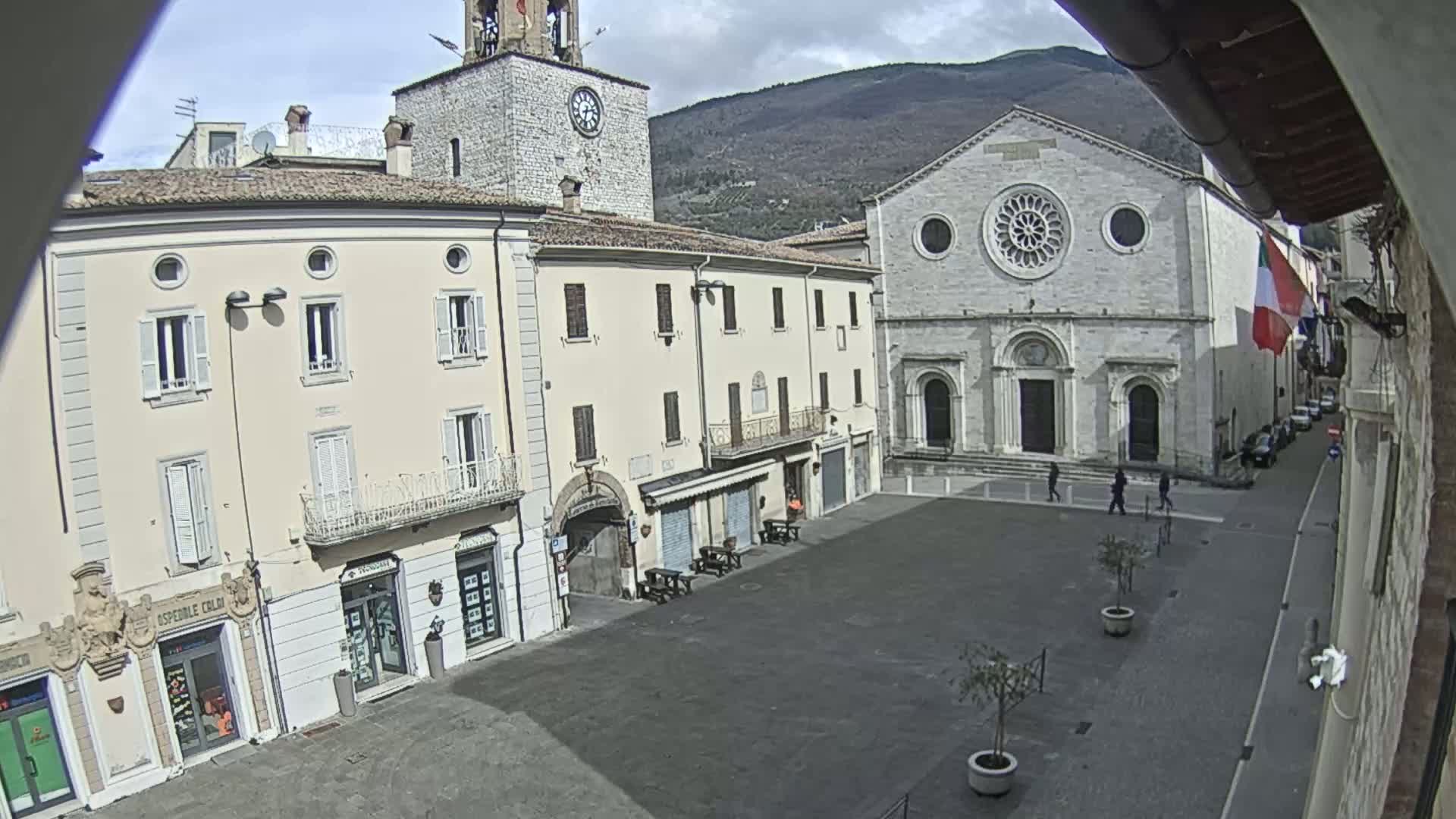 A mostly empty town square with several stone buildings, including a church with a rose window, under a partly cloudy sky.