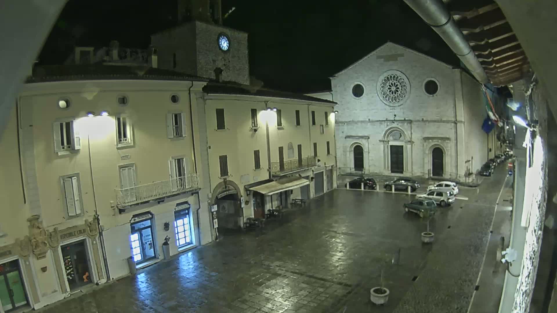 A wet, nighttime view of a town square with several buildings, including a church with a rose window, and a few parked cars.