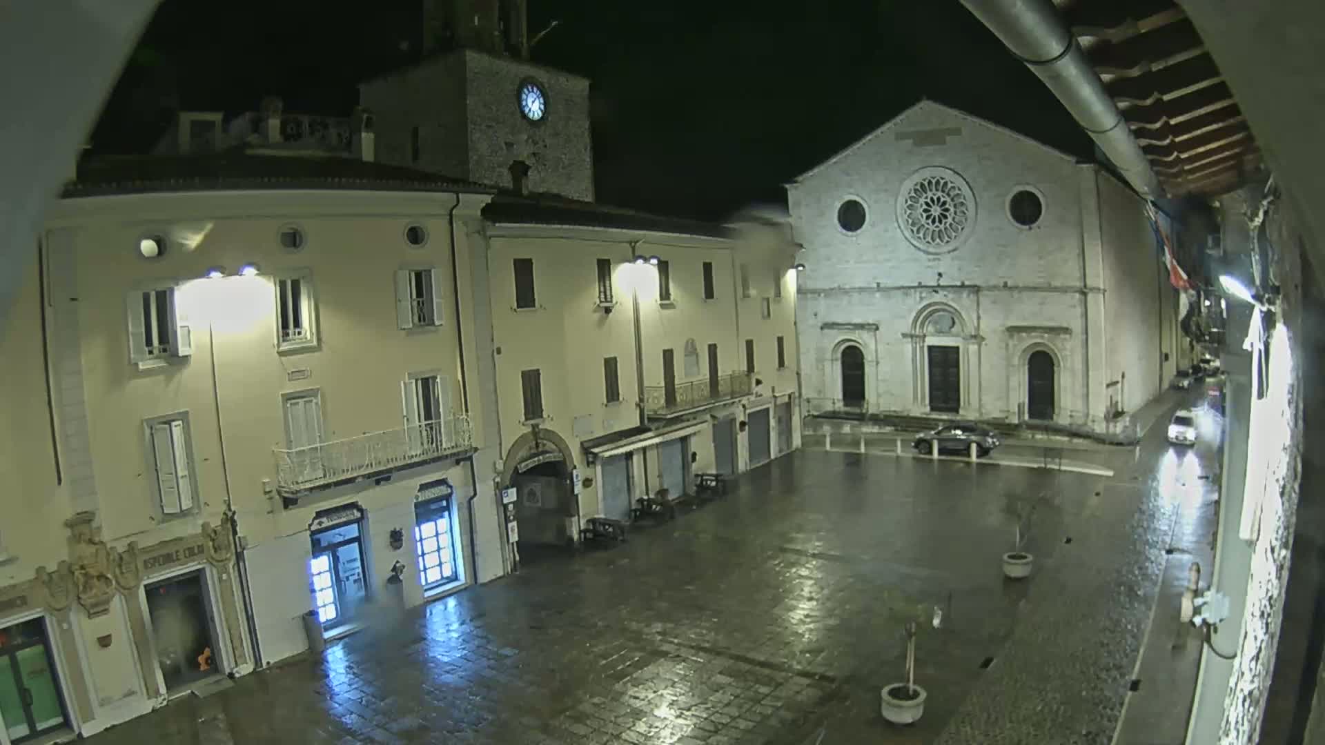 A rain-slicked town square at night, featuring several buildings, including one with a clock tower and another with a large rose window.