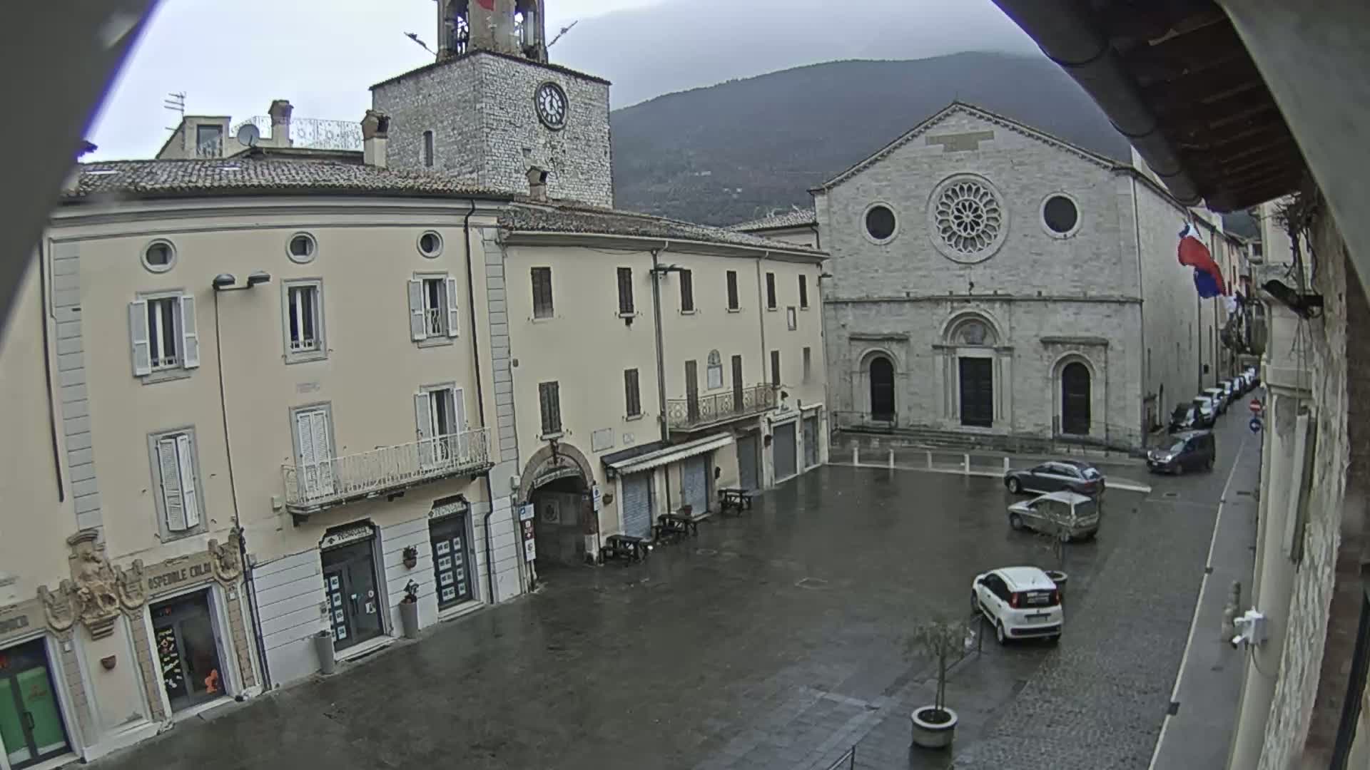A town square with several buildings, including a clock tower and a church, is shown on a rainy day, with a few cars parked in the square.