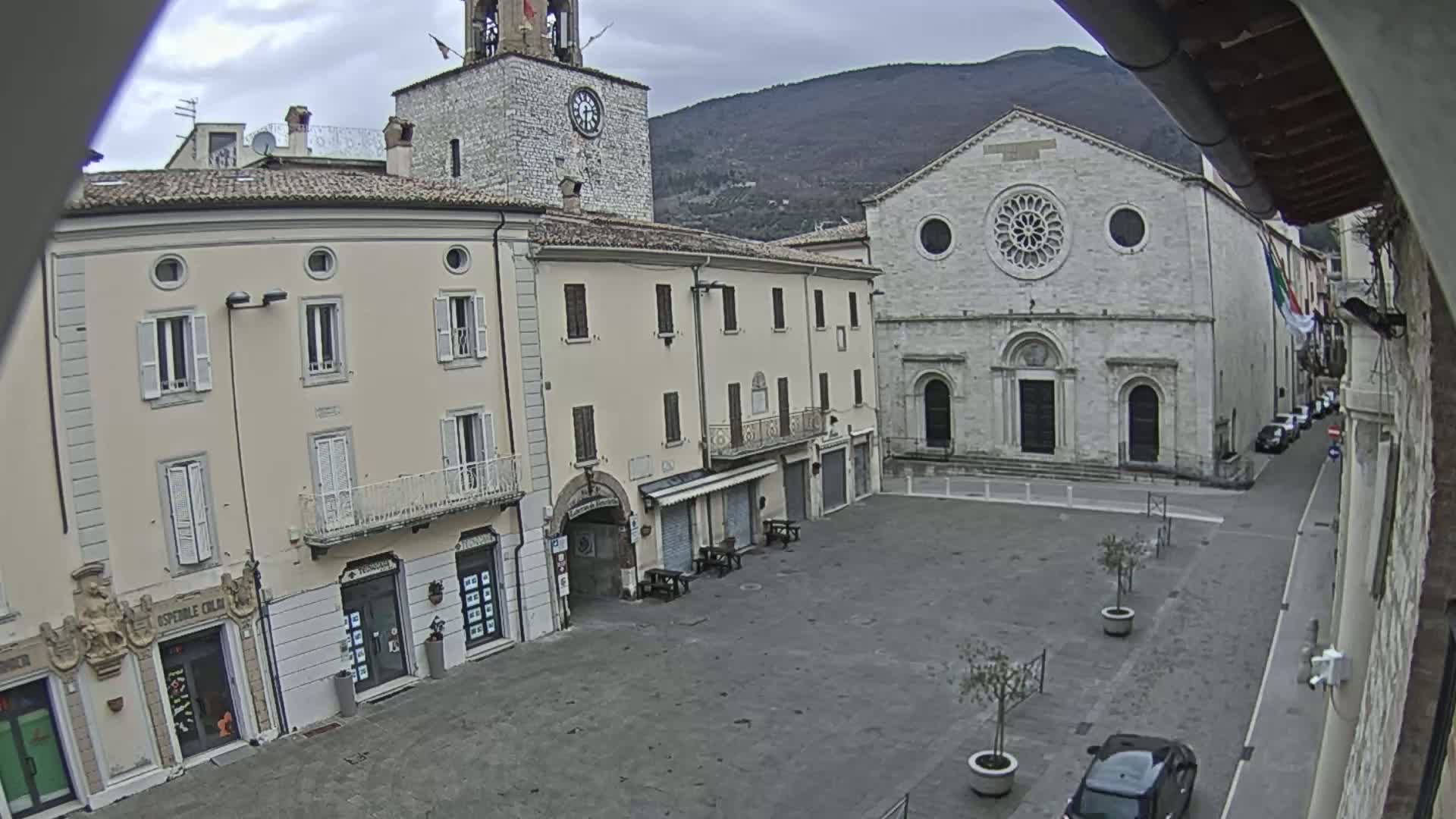 A mostly empty town square, under an overcast sky, is surrounded by aged stone buildings, including a clock tower and a church with a rose window.