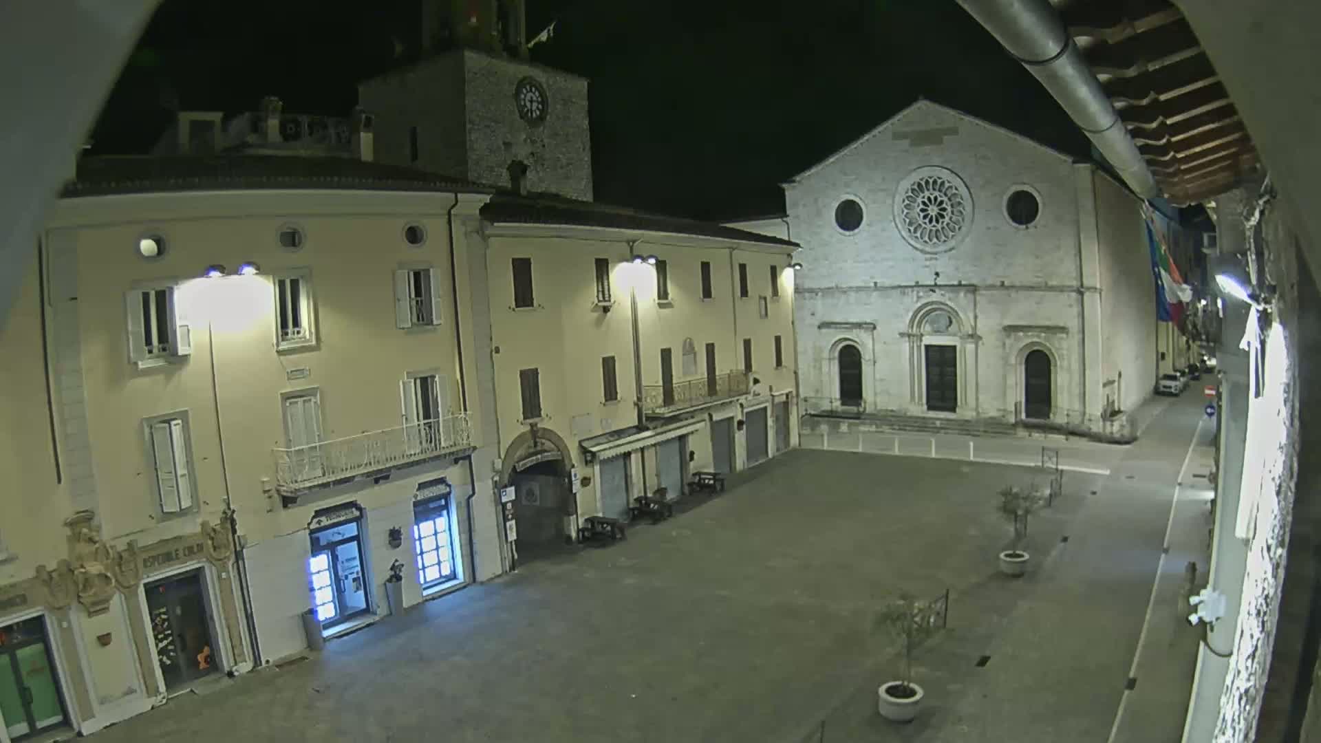 A nighttime view of a town square, featuring buildings with illuminated shopfronts, a church with a rose window, and sparsely planted trees.