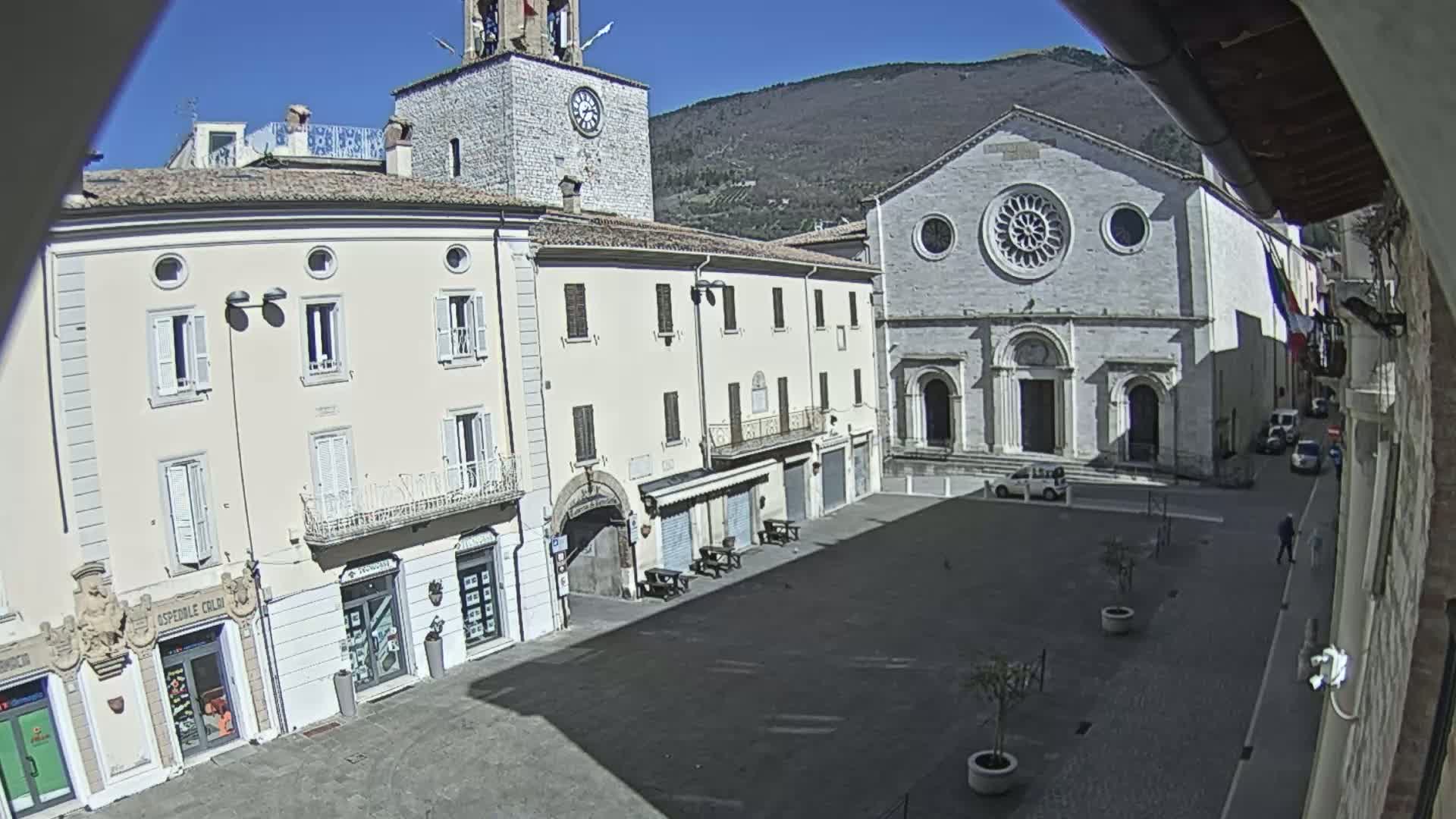 A sunny town square is viewed from above, showing several light-colored buildings, including a church with a rose window, a clock tower, and shops, under a clear blue sky and hills in the background.